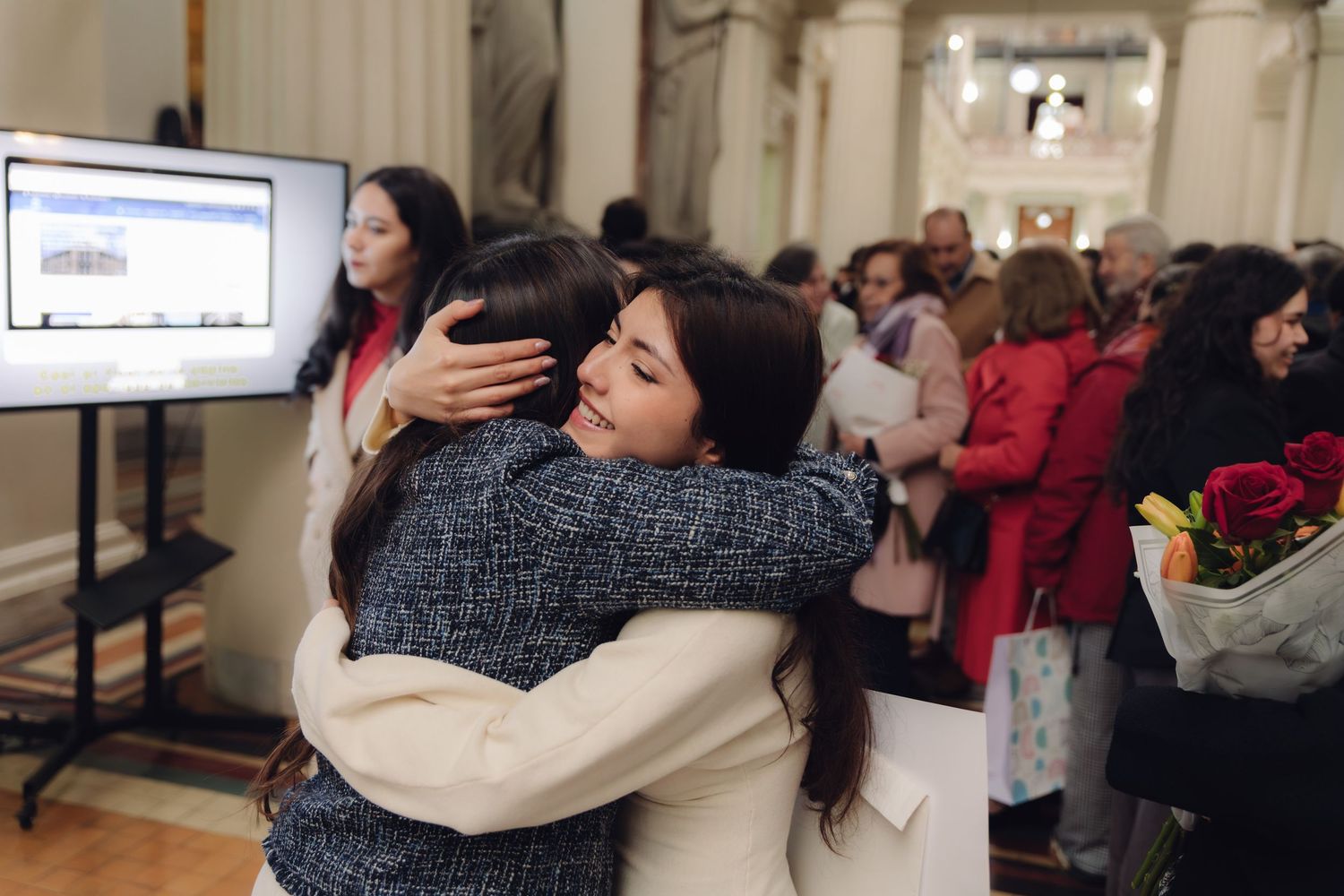 Two graduates embrace emotionally at indoor graduation celebration ceremony.