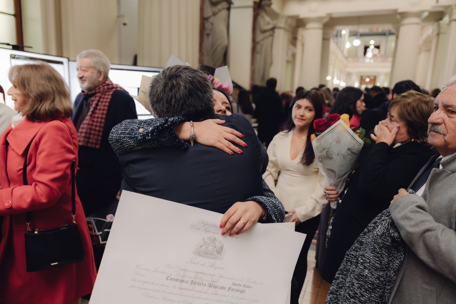 Graduate hugs attendee while proudly holding diploma certificate at graduation ceremony.
