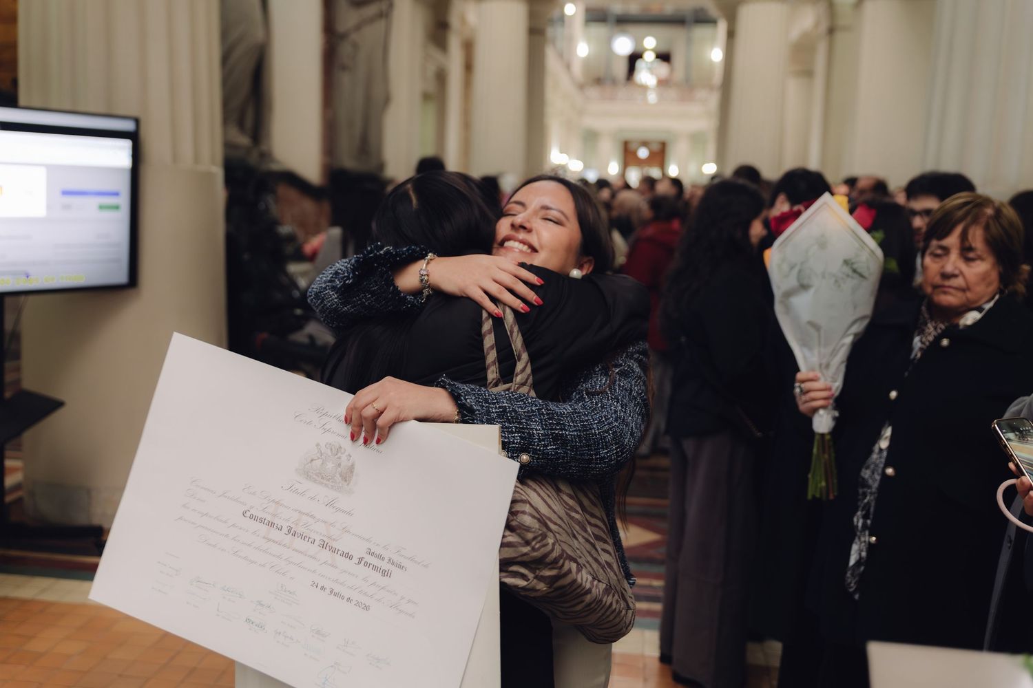 Emotional graduate hugs family member while holding official diploma at graduation event.