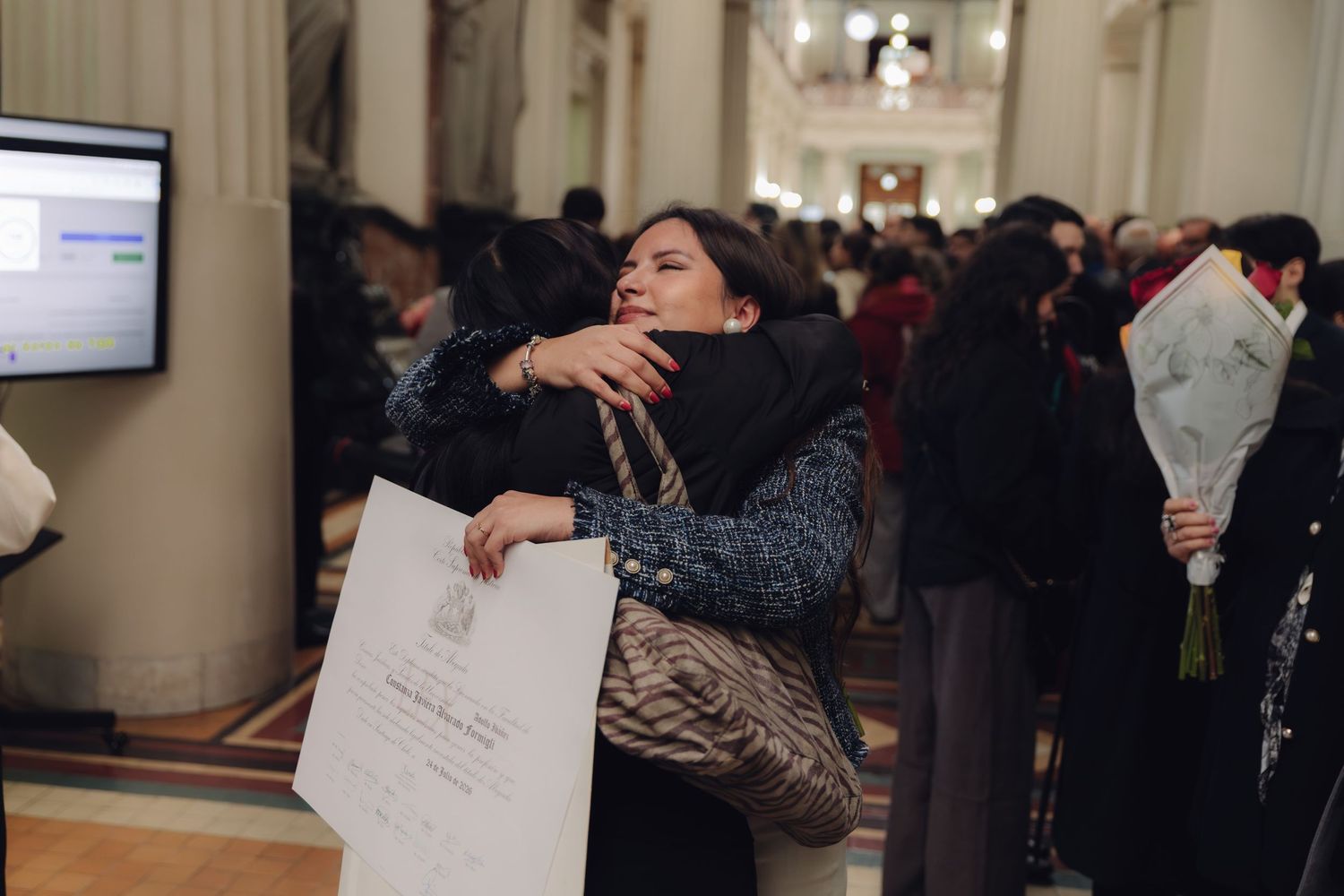 Woman hugging someone while holding a diploma certificate at a graduation ceremony indoor venue.