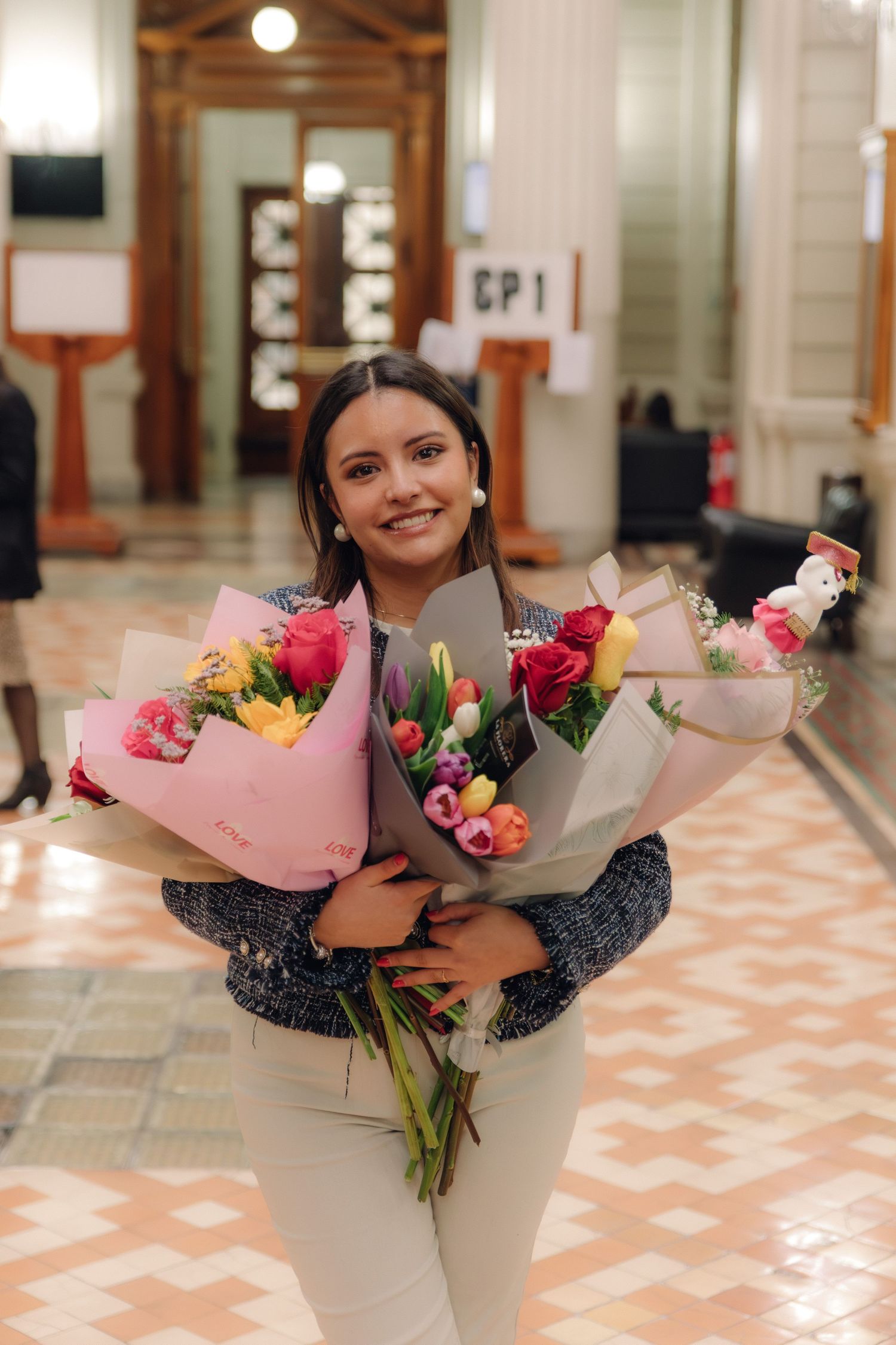 Smiling young woman holding three large colorful flower bouquets in an elegant indoor hall.