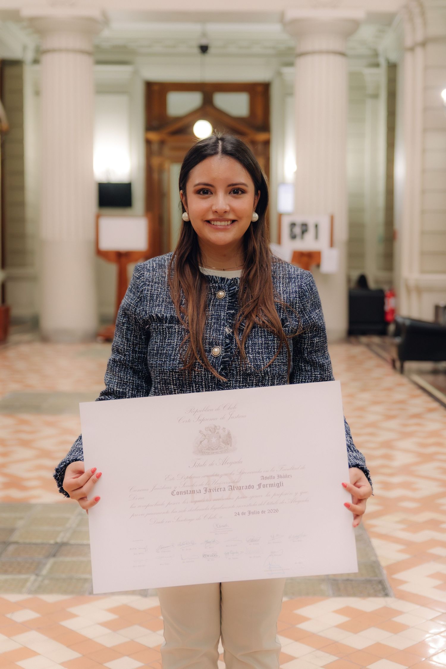 Smiling young woman in blue tweed jacket proudly displaying her graduation diploma certificate indoors.