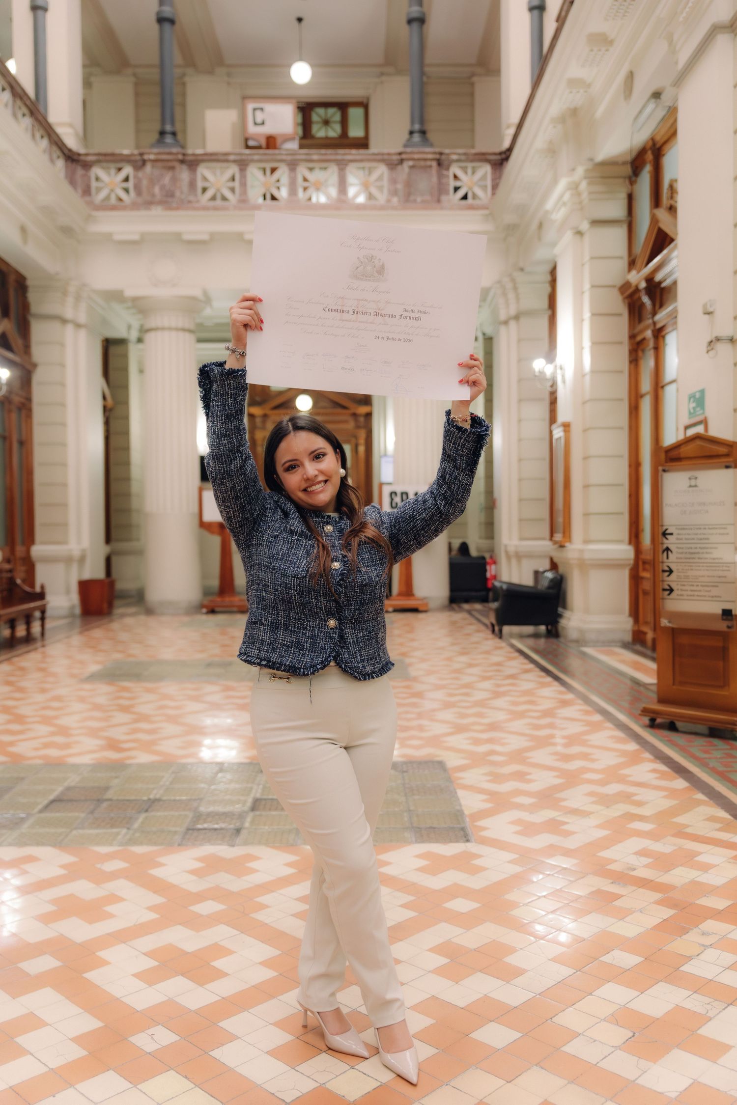 Joyful young woman raising graduation diploma above her head in a grand ornate building hall.