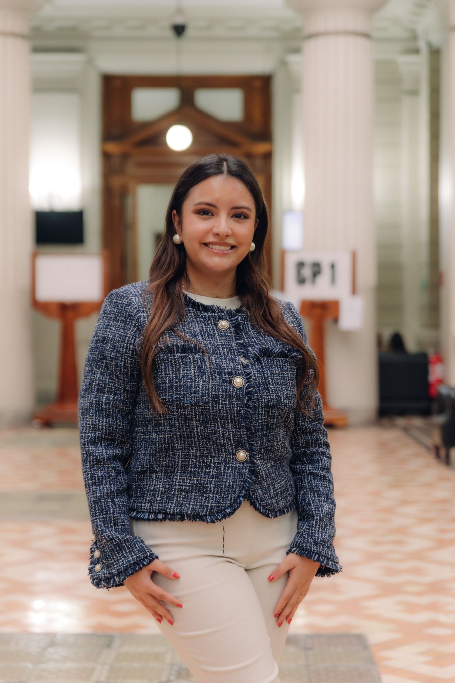 Young woman in blue tweed jacket smiling confidently in an elegant columned interior hallway.