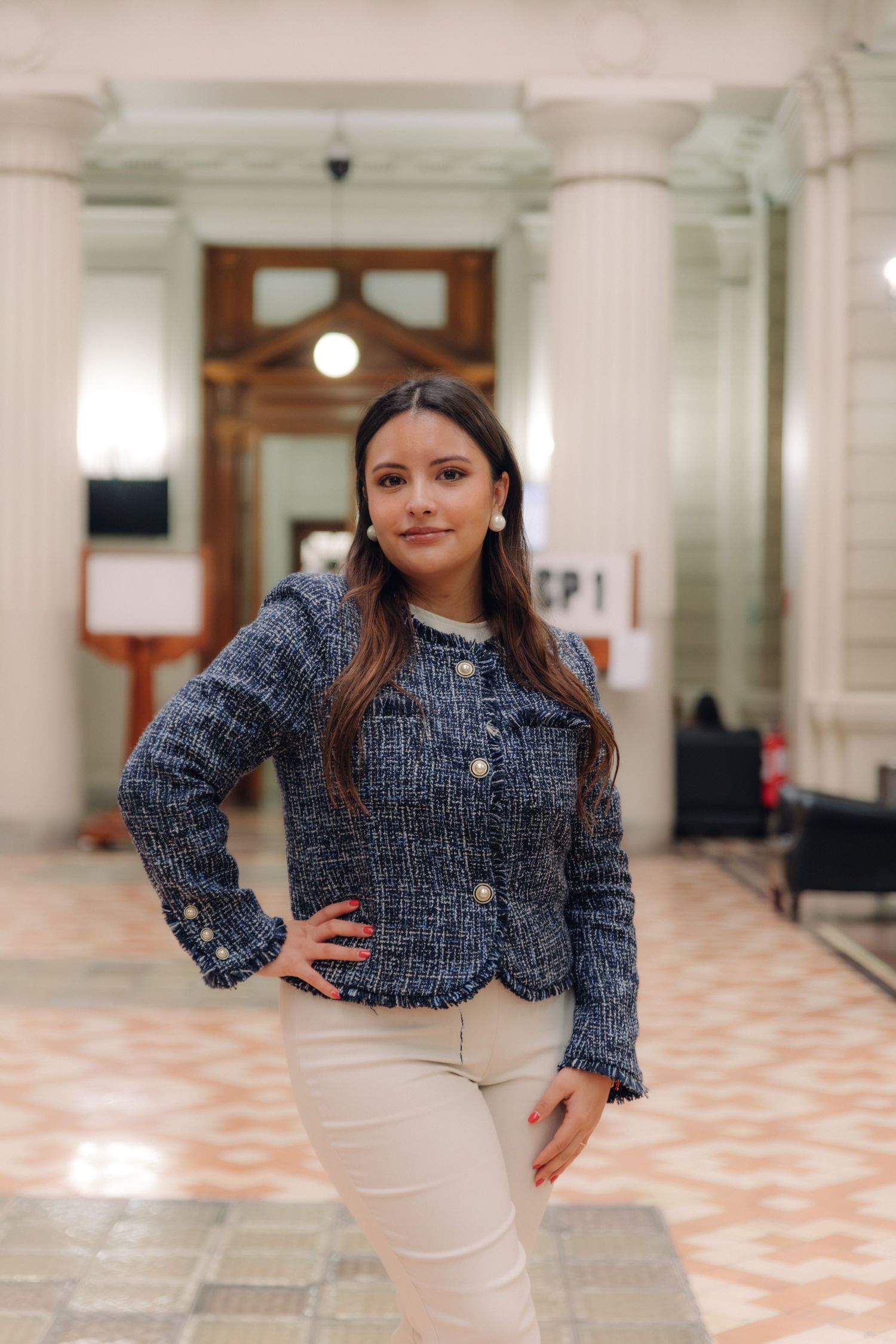 Young woman in blue tweed jacket posing with hand on hip in a grand interior hall.