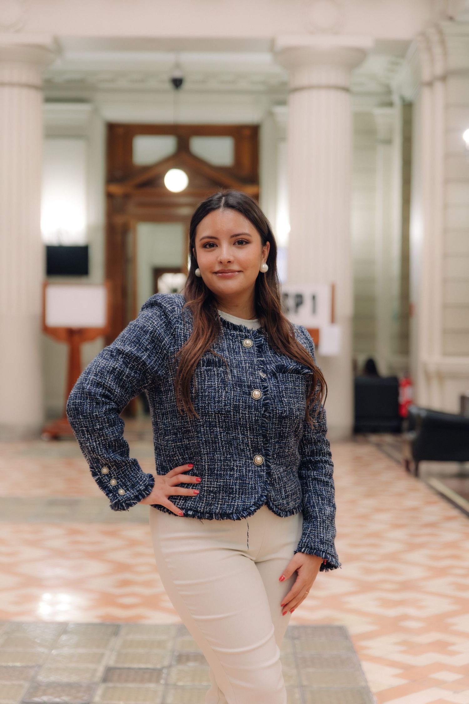 Smiling young woman in blue tweed jacket posing confidently in an elegant columned building interior.