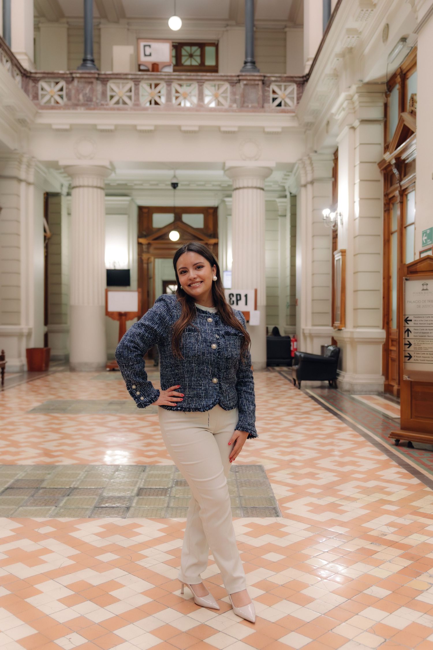 Young woman in blue tweed jacket posing in a grand ornate hall with geometric tiled floor.