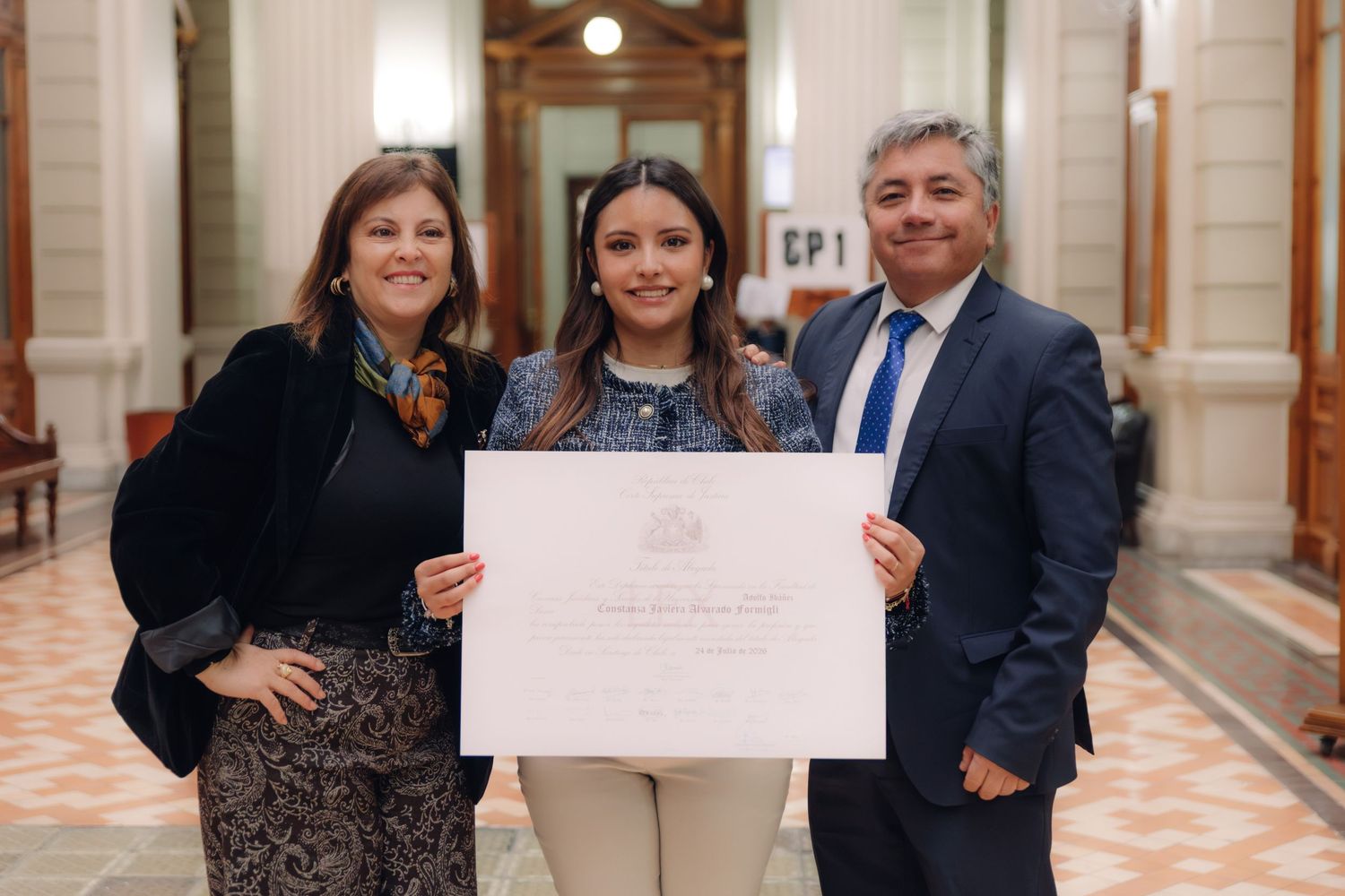 Young woman holds diploma flanked by proud parents in elegant hall.