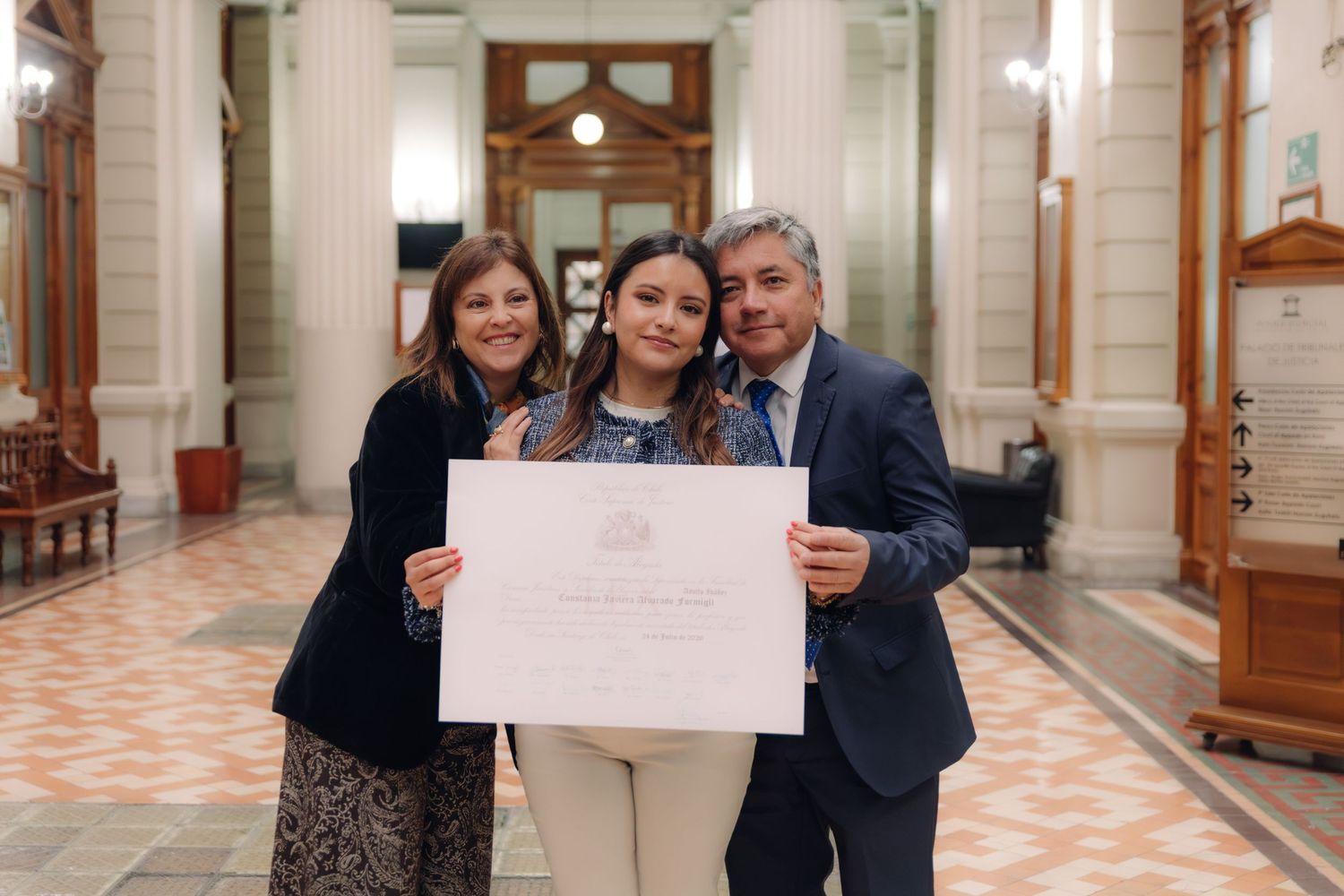 Graduate poses with certificate between smiling parents indoors.