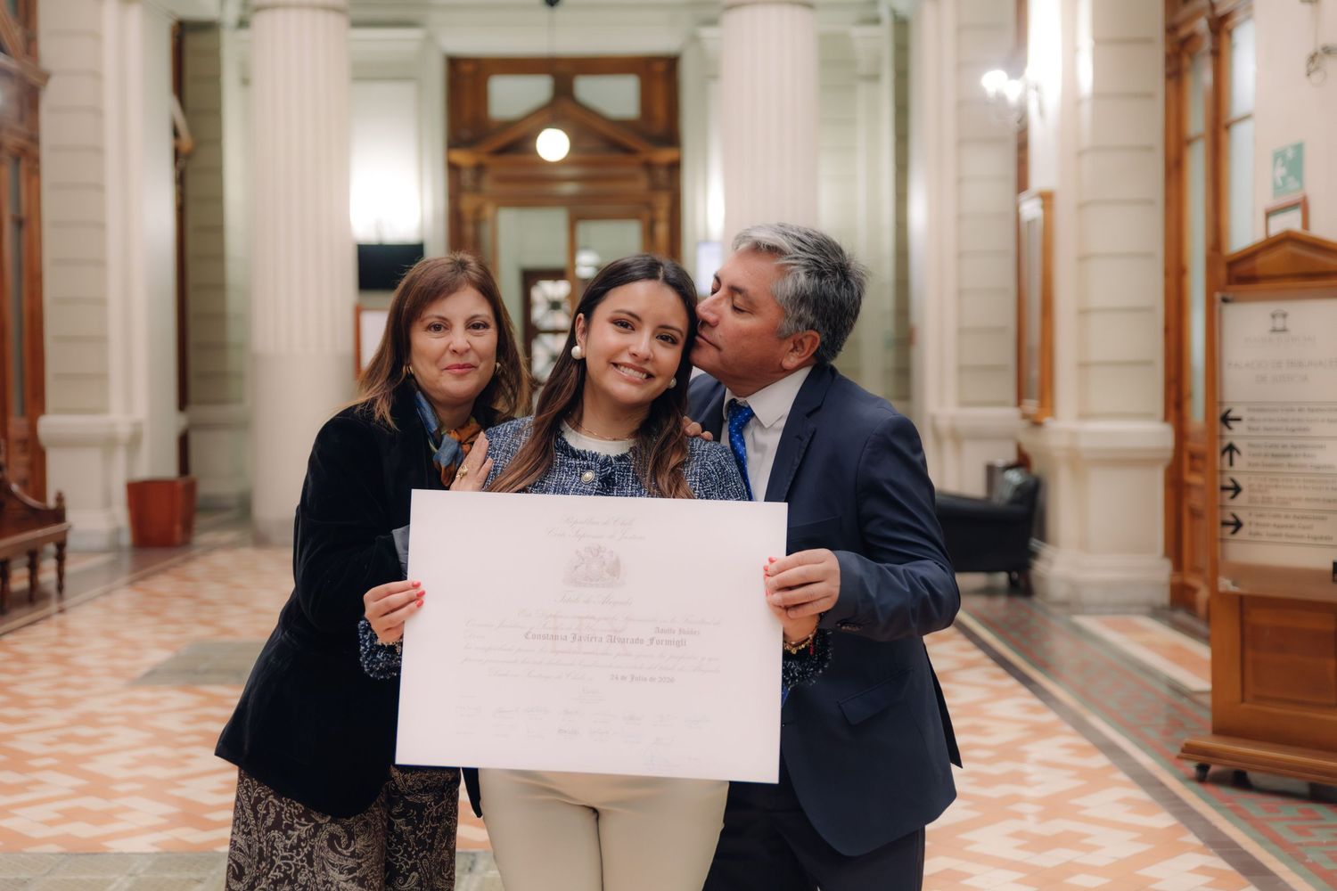 Man kisses graduate on cheek as woman smiles beside diploma.