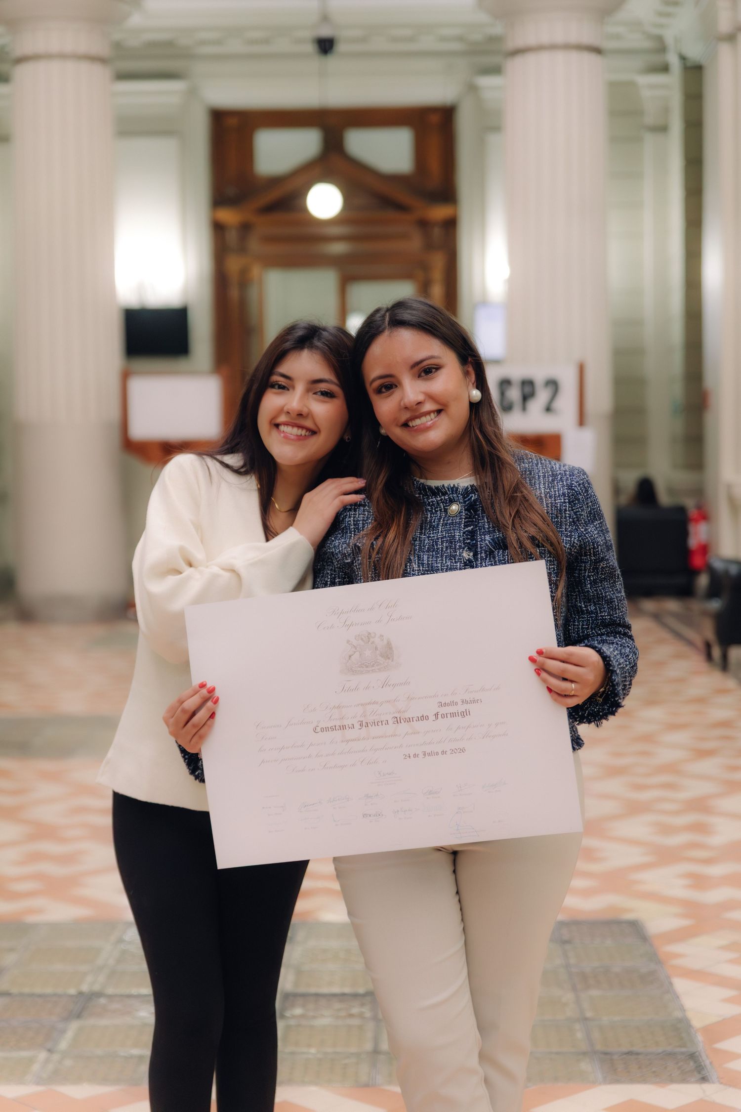 Two young women smile holding graduation certificate in grand hall.