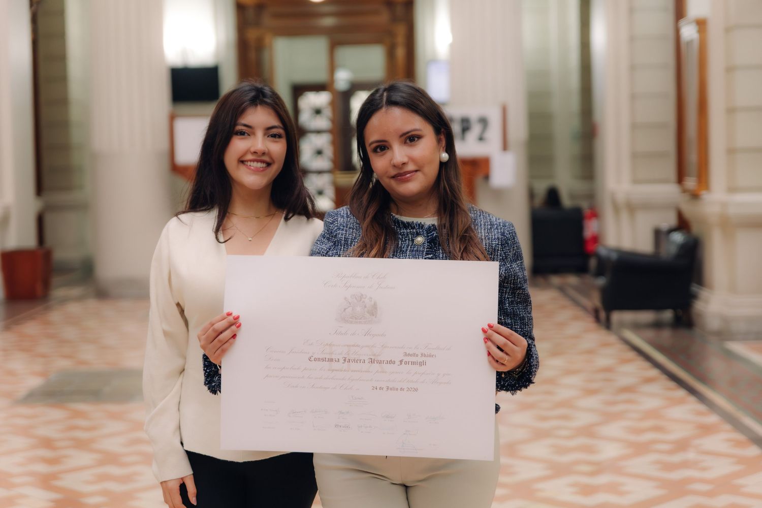 Two women pose together holding official diploma in corridor.
