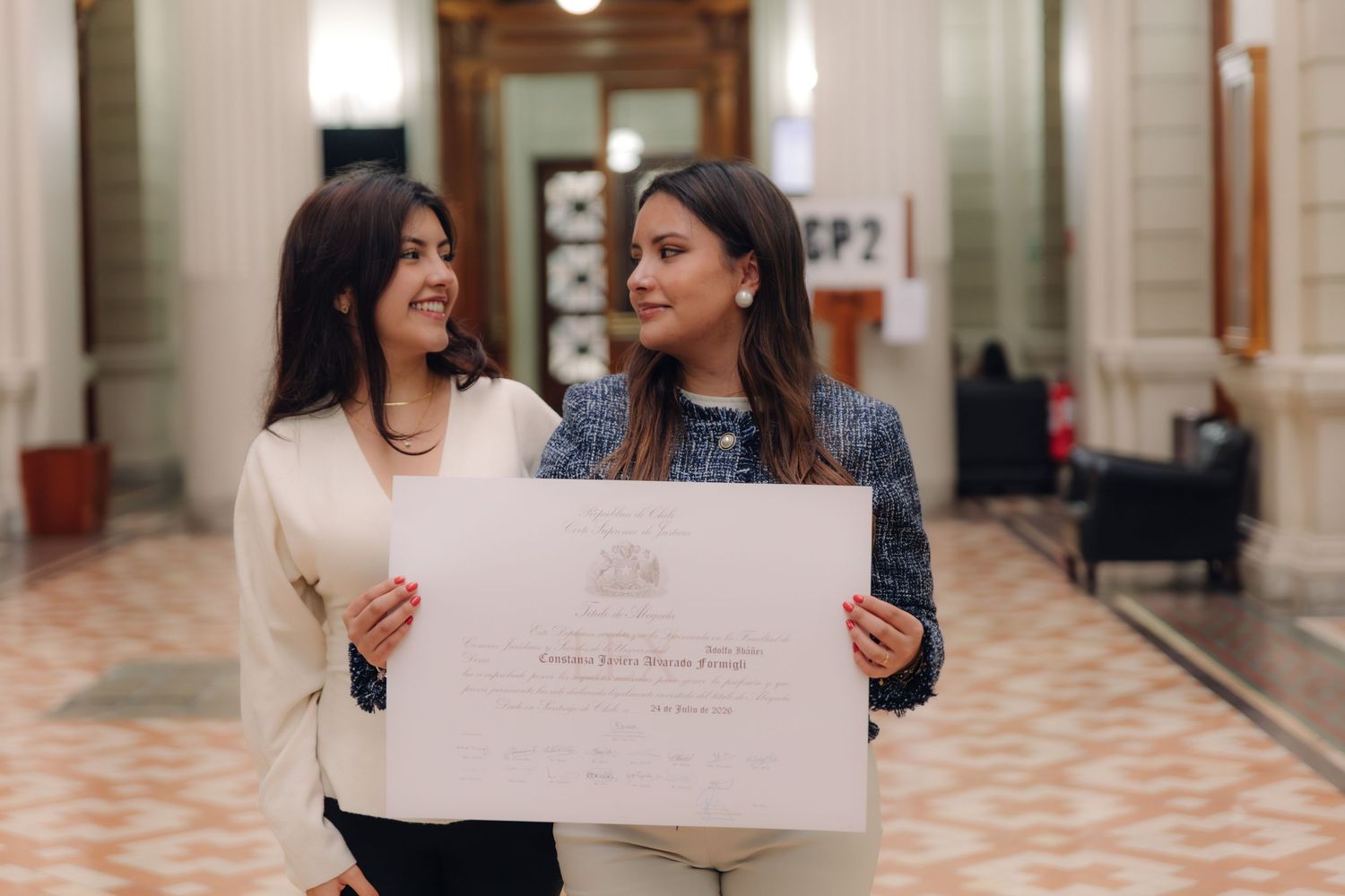 Two smiling women share a look while holding graduation certificate.
