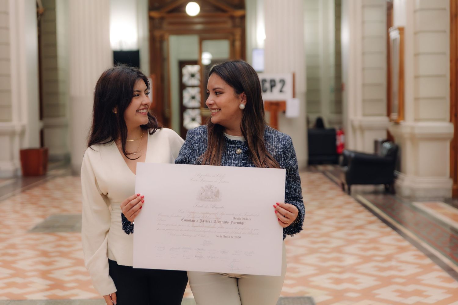 Two women laugh together while displaying official diploma indoors.