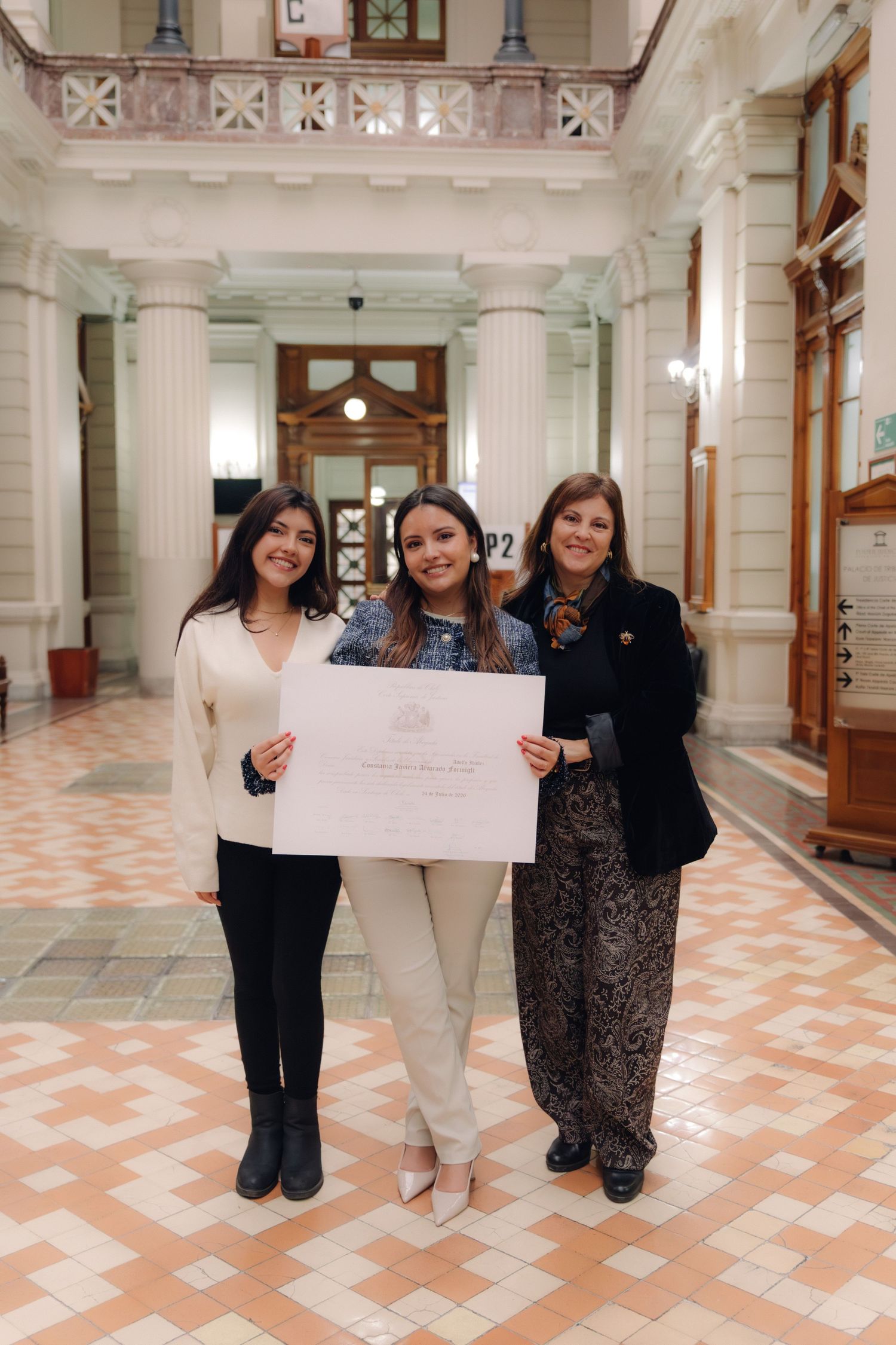 Three women proudly display a certificate in an elegant columned hallway.