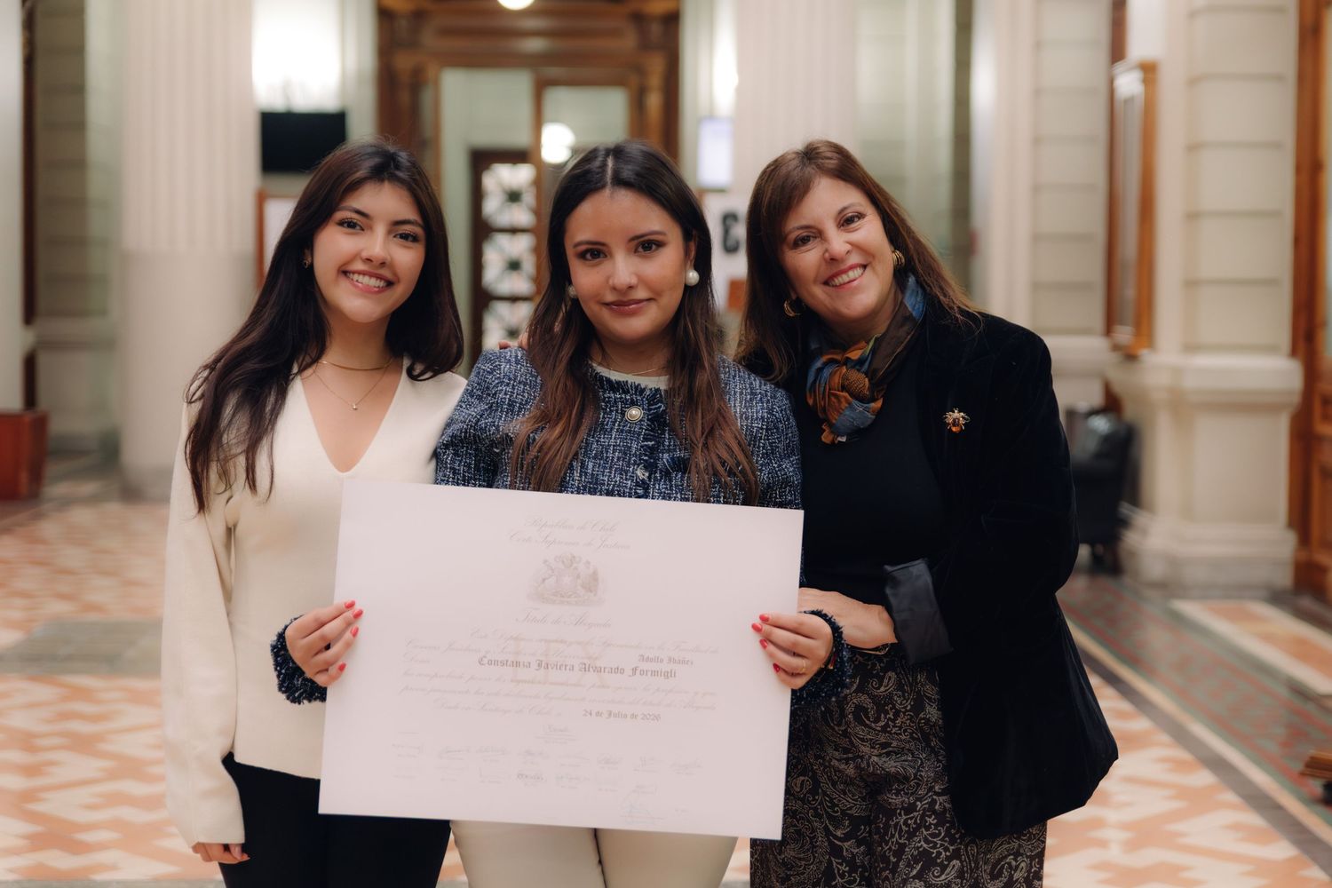 Three smiling women pose together holding a formal certificate indoors.