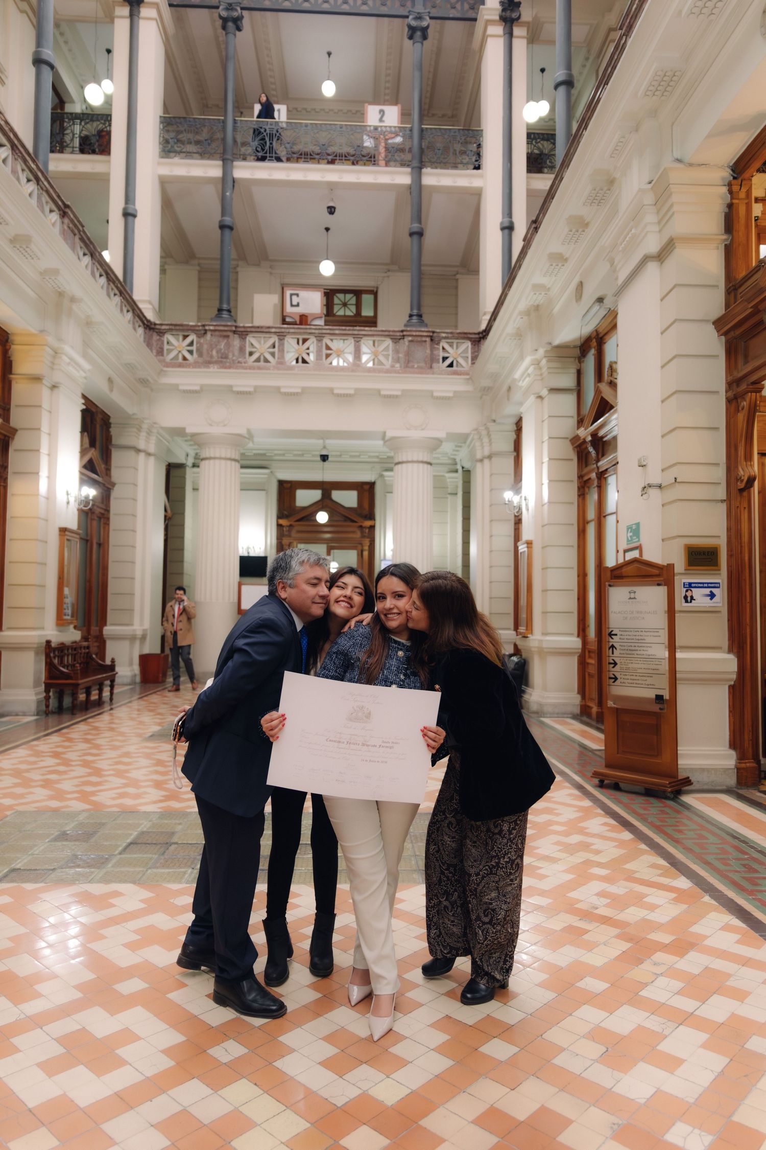 Three people share a joyful embrace holding a certificate in a hall.