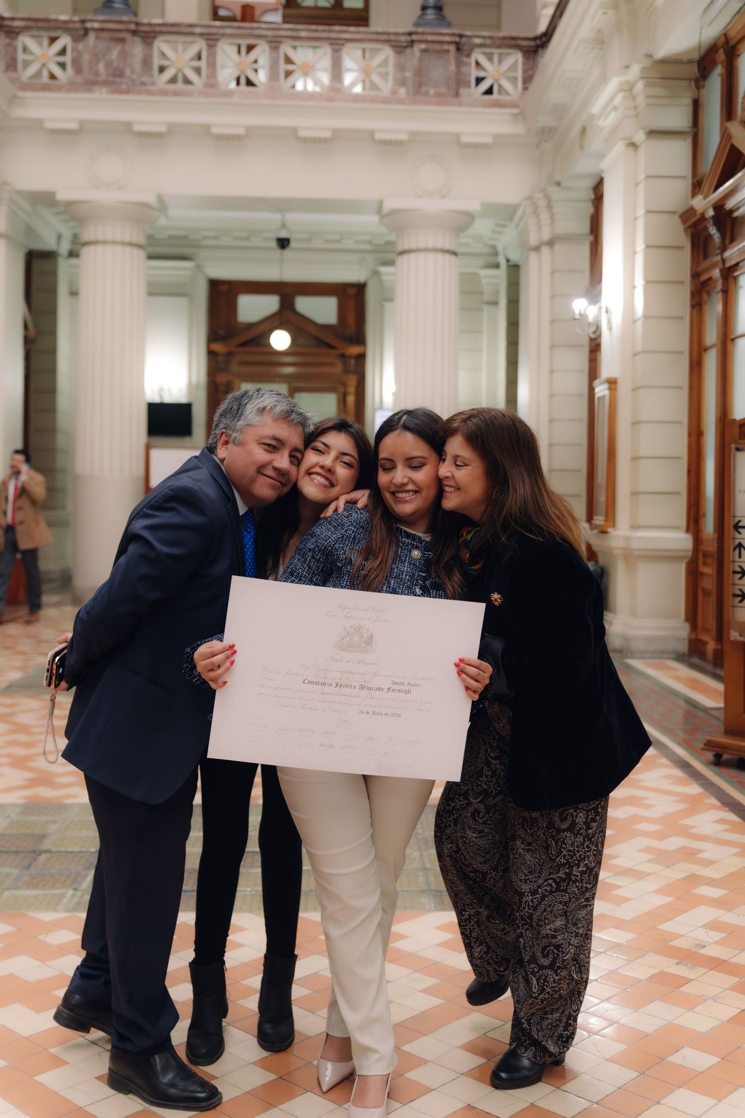 Family of four celebrates with hugs holding a certificate in a hall.