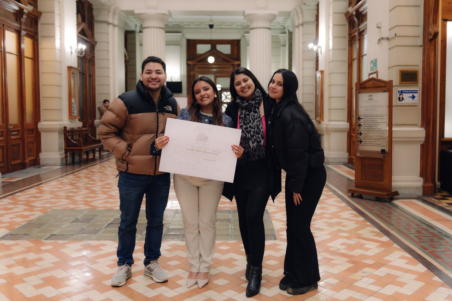 Four young people smile holding a certificate in an elegant corridor.