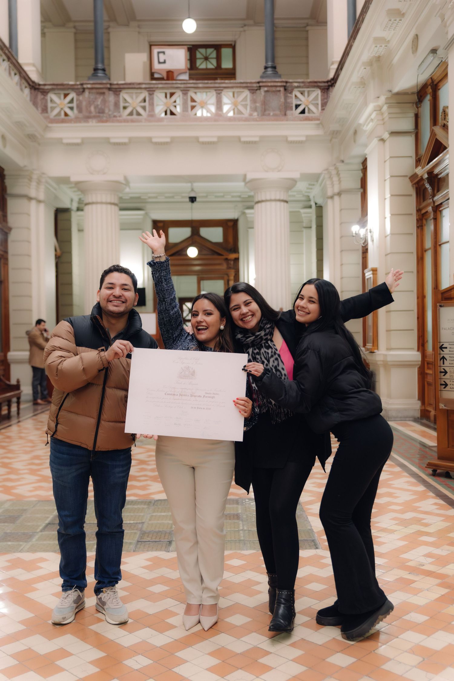 Four celebrating friends wave and hold a certificate in a grand hall.