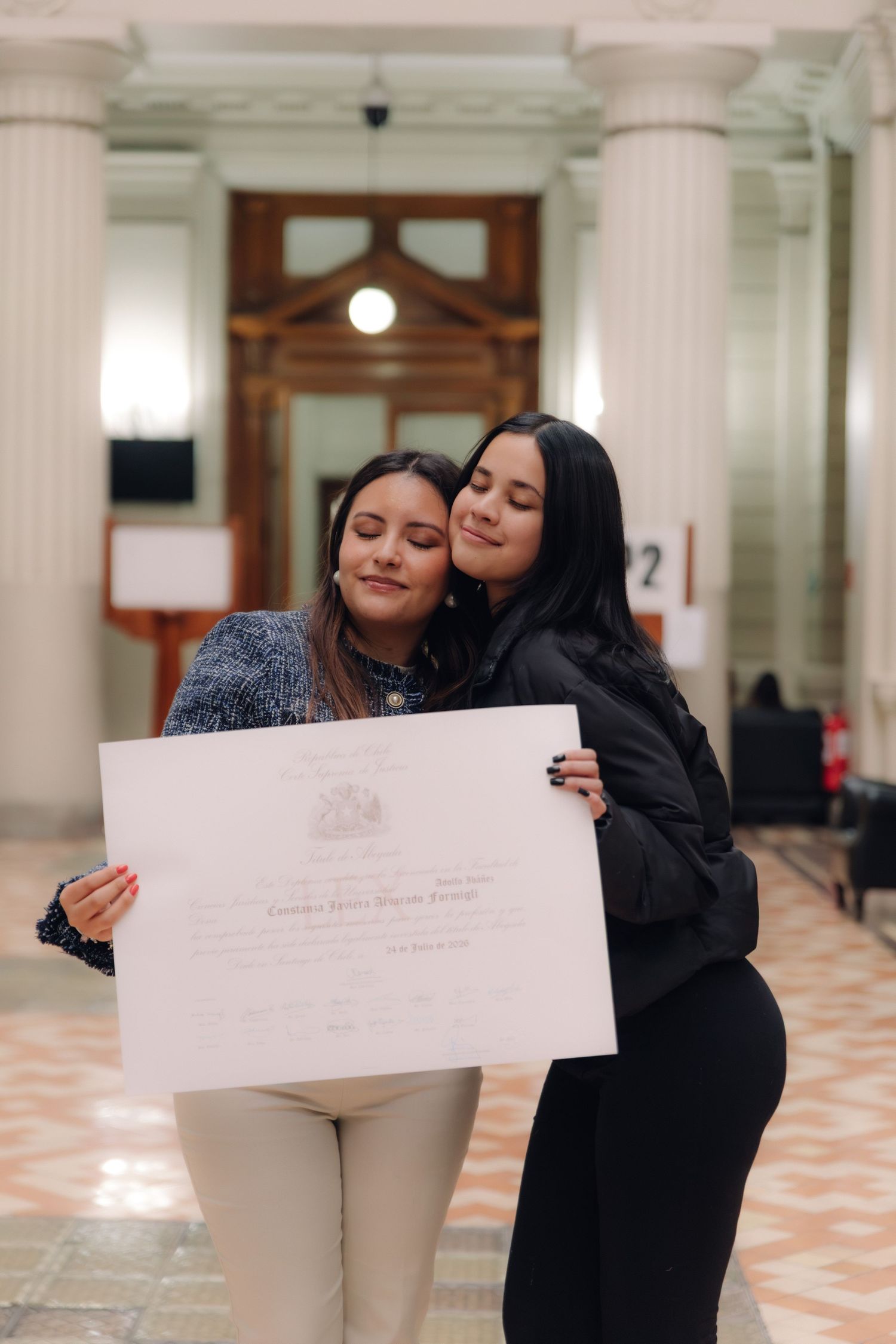 Two young women proudly holding a diploma certificate together in an elegant columned hall.