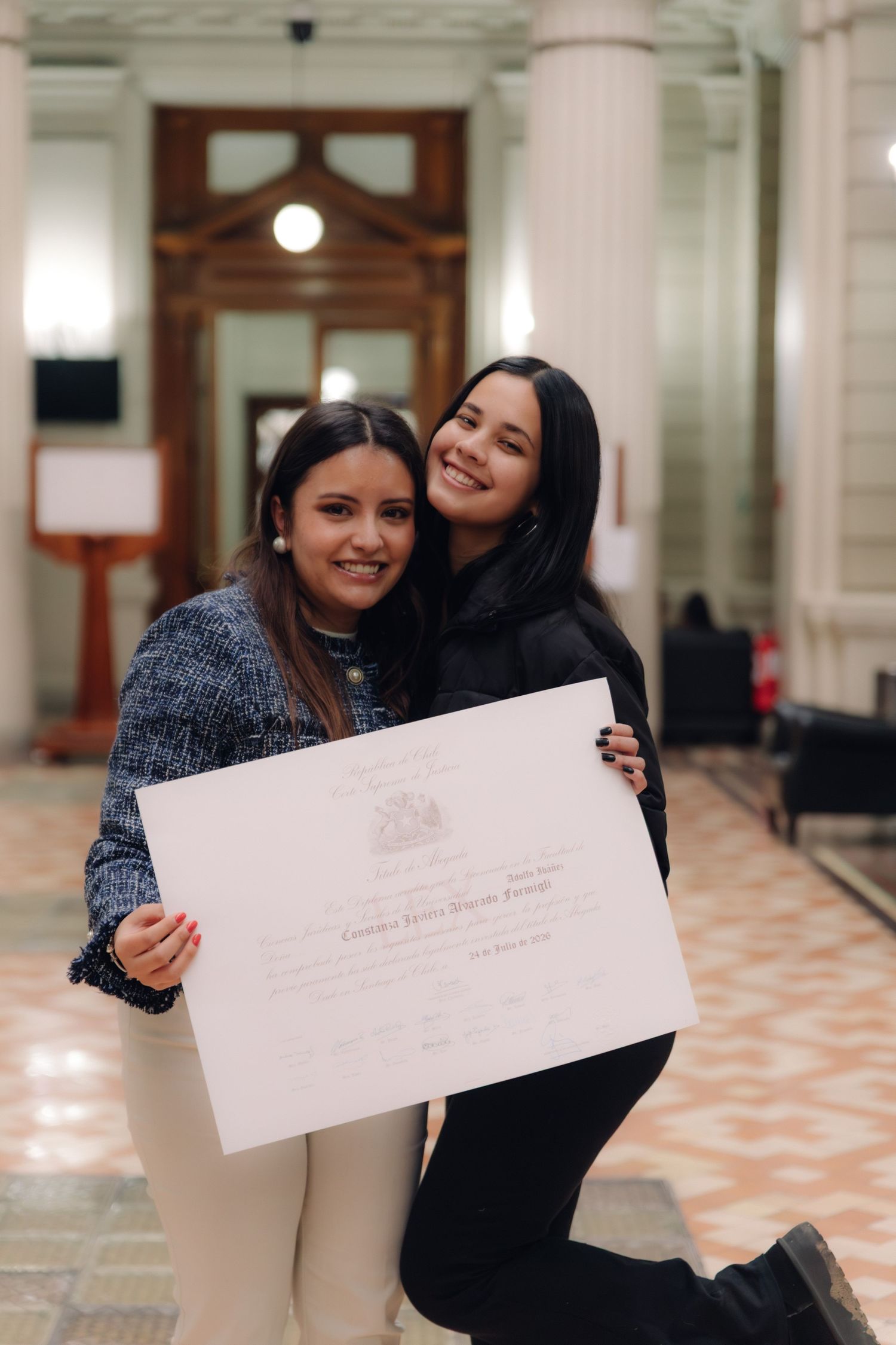 Two smiling women posing with a graduation diploma in a grand academic building interior.