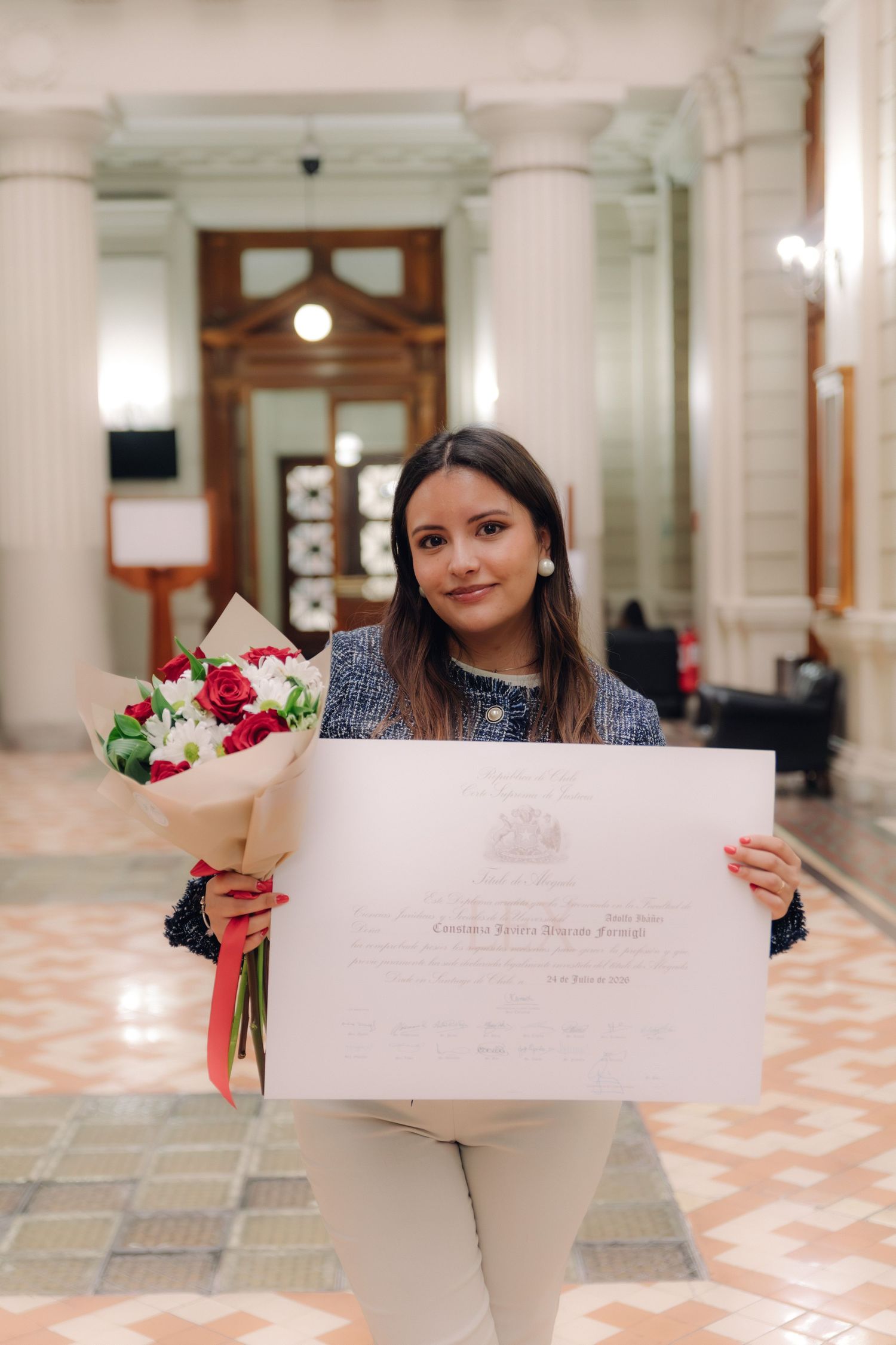 Young woman holding a diploma and red rose bouquet during graduation ceremony in historic hall.