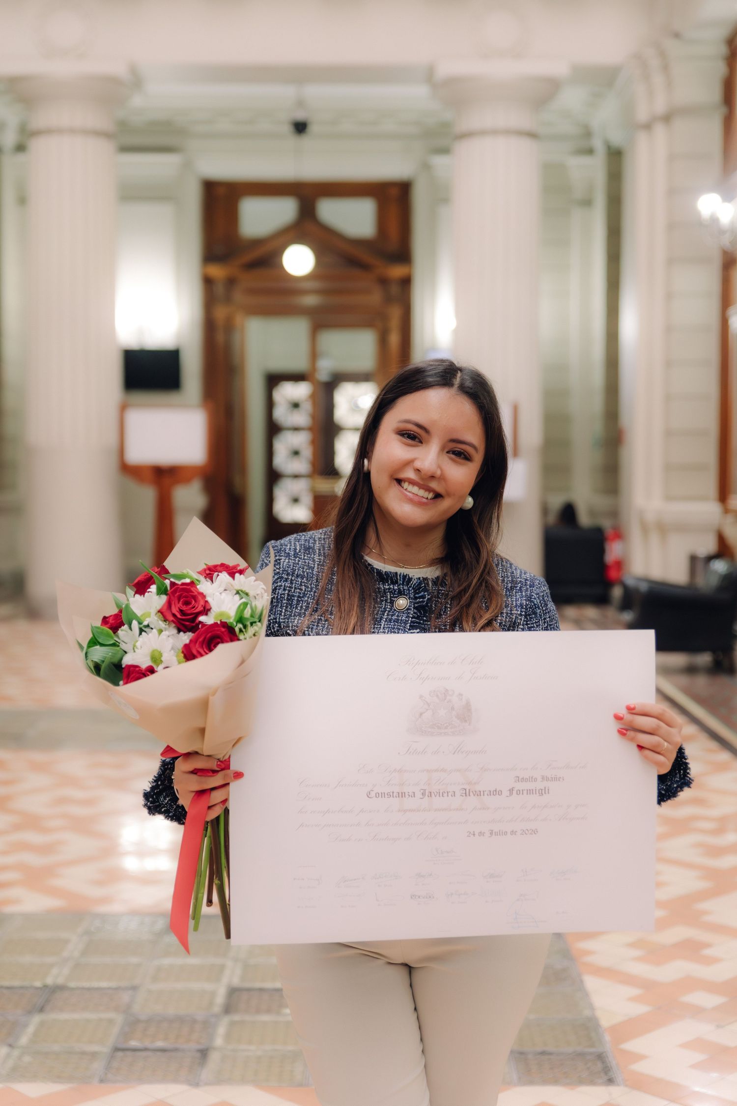 Smiling graduate proudly displaying diploma and flower bouquet in elegant columned building.