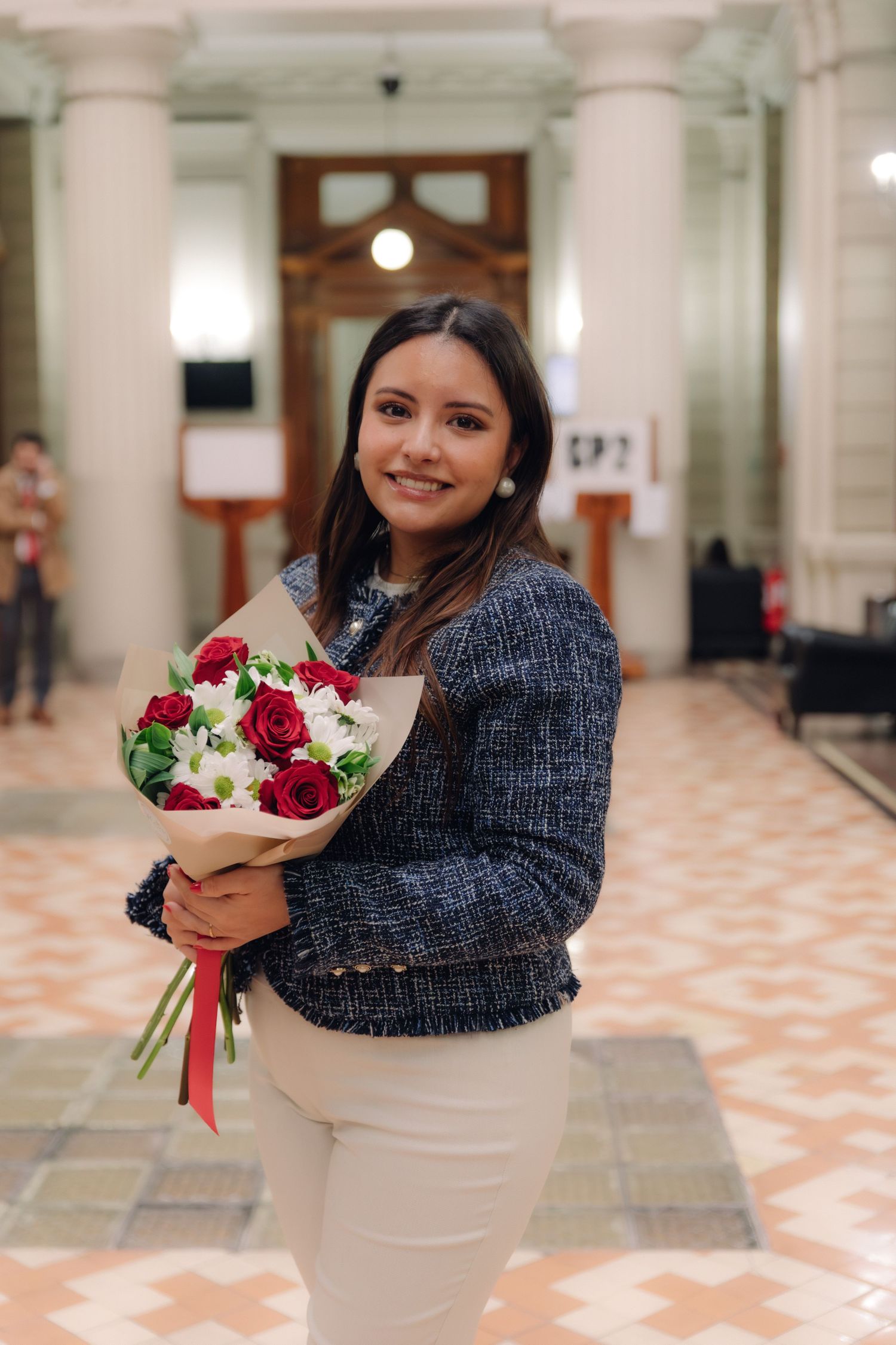 Young woman in blue tweed jacket holding red roses and white flower bouquet at graduation.