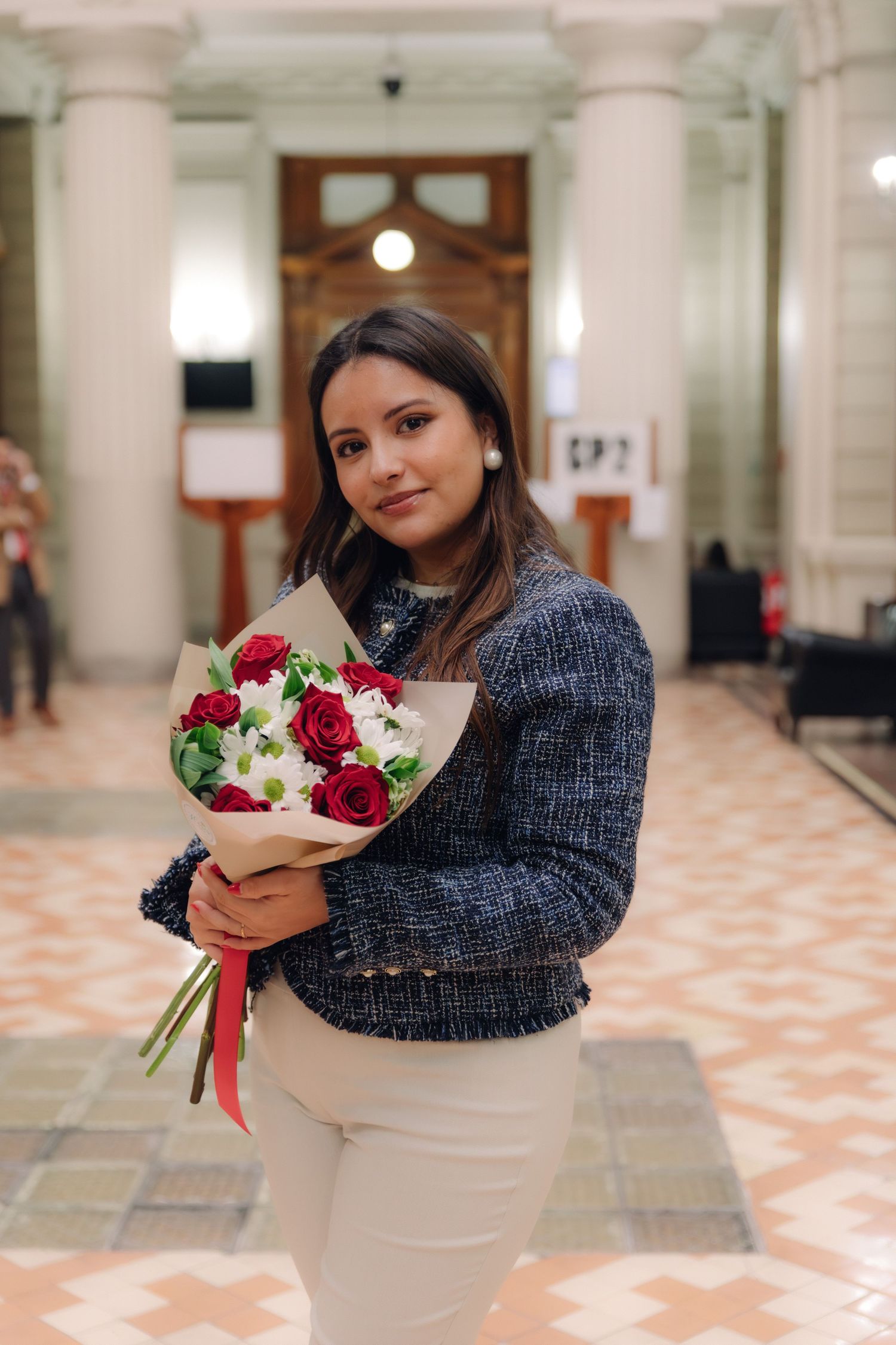 Graduate in blue tweed blazer holding red and white flower bouquet in ornate building hall.