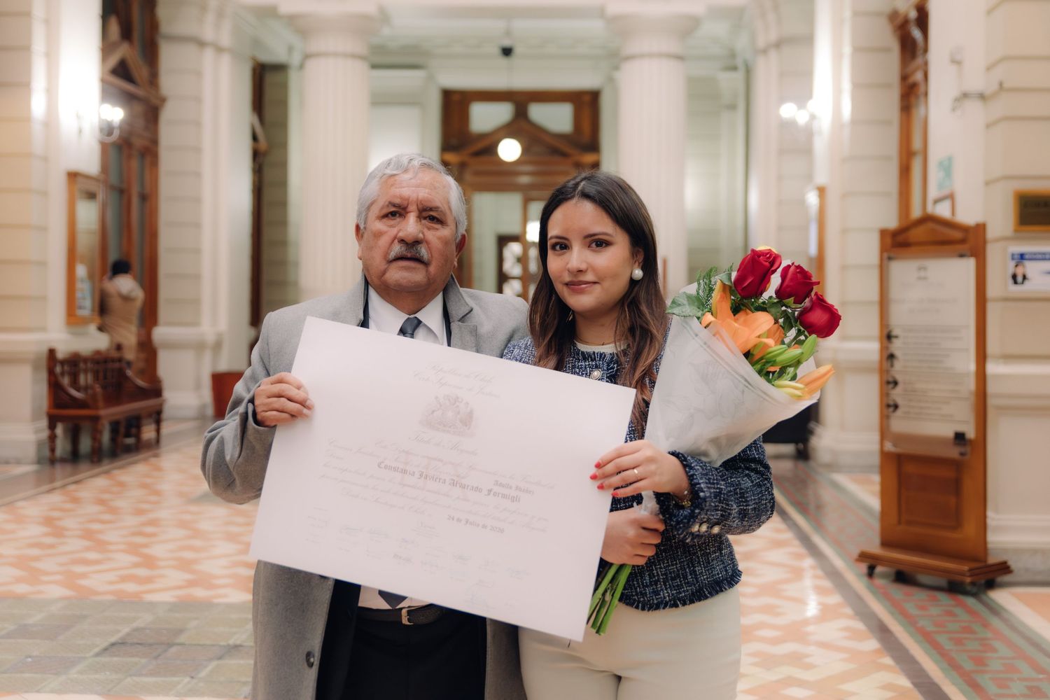 Elderly man and young female graduate holding diploma with flower bouquet in historic hall.