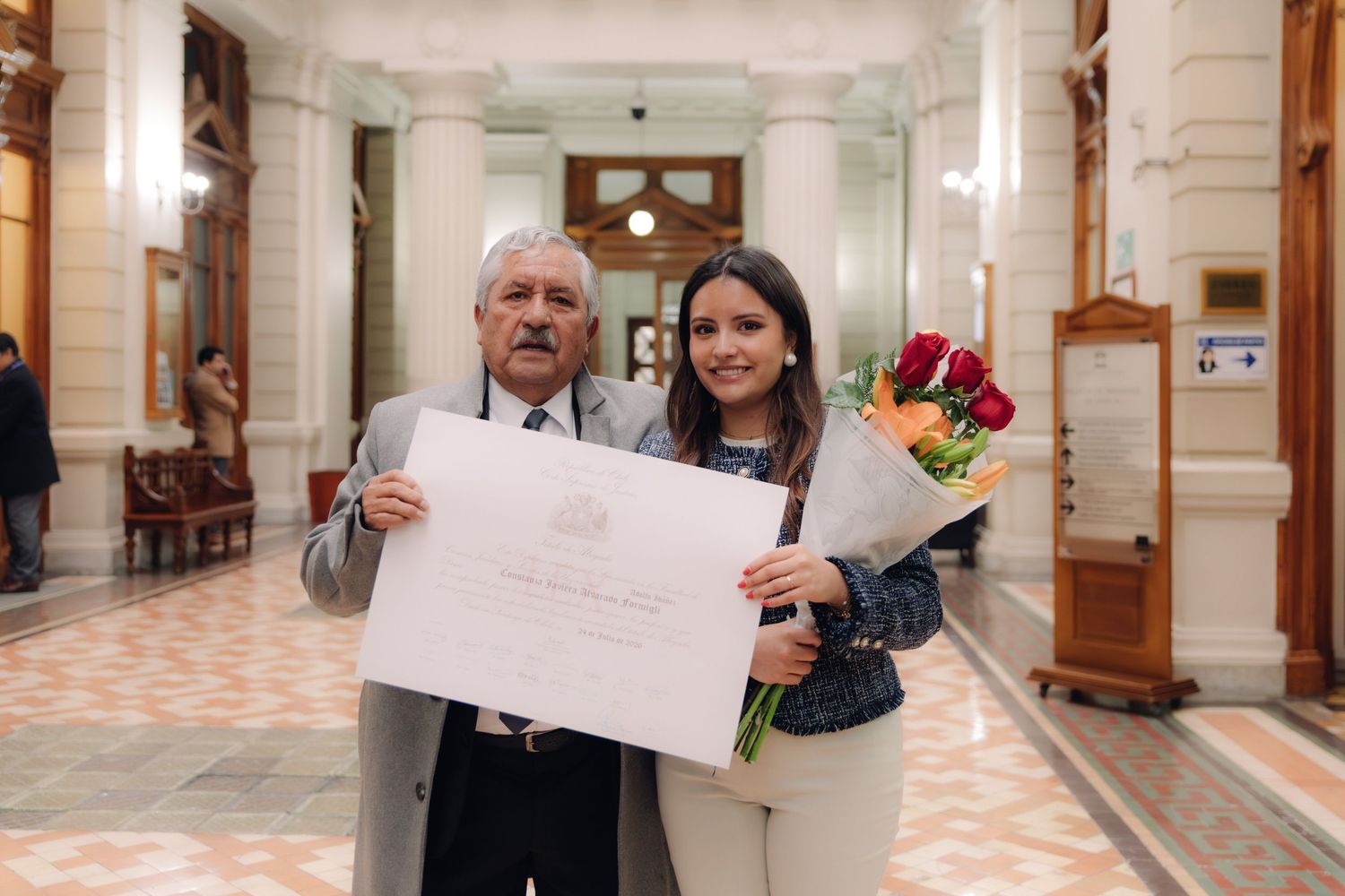 Senior man and young woman posing with graduation diploma and colorful bouquet indoors.