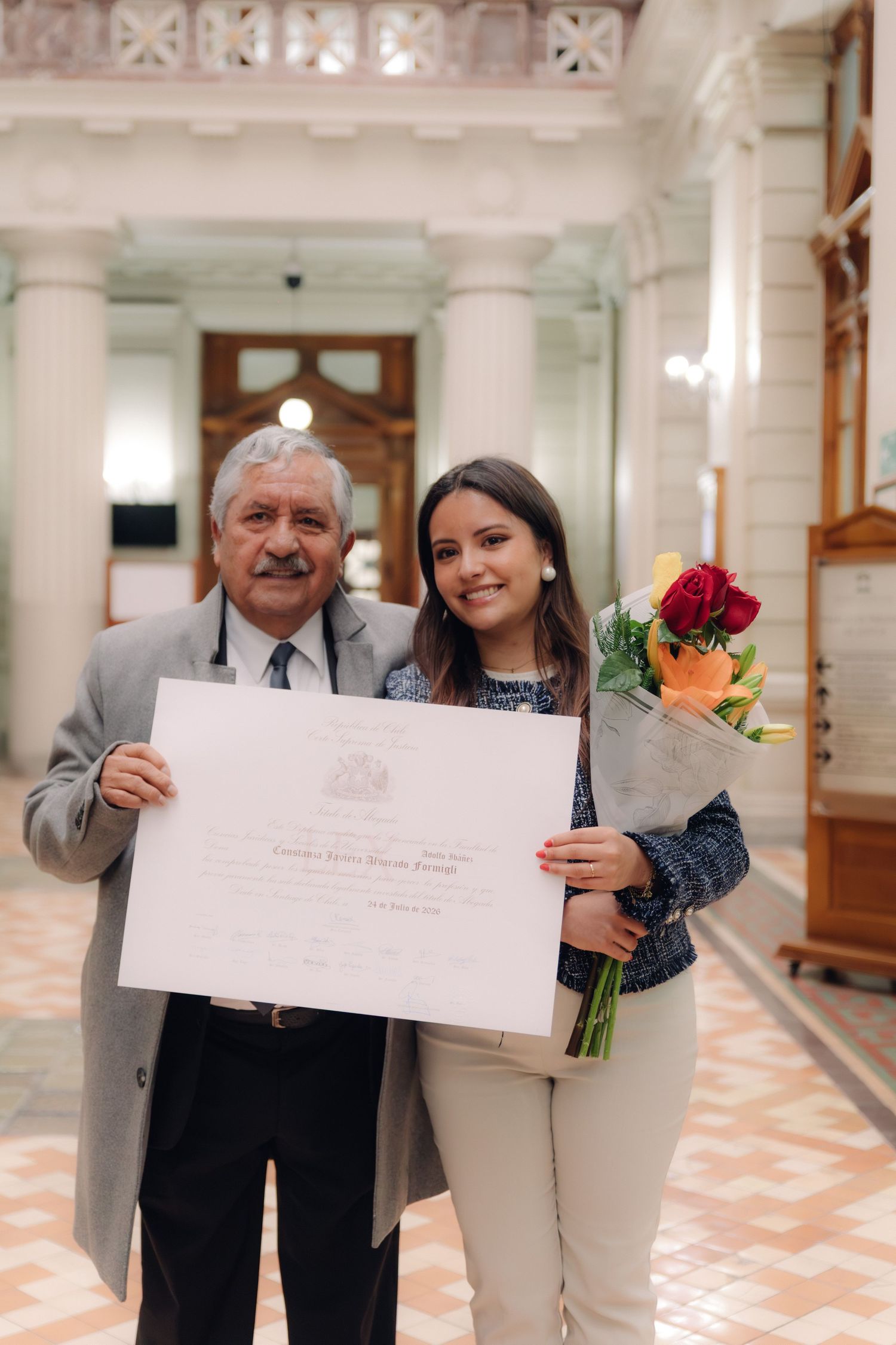 Older man and young woman proudly display an official diploma certificate indoors with flowers.