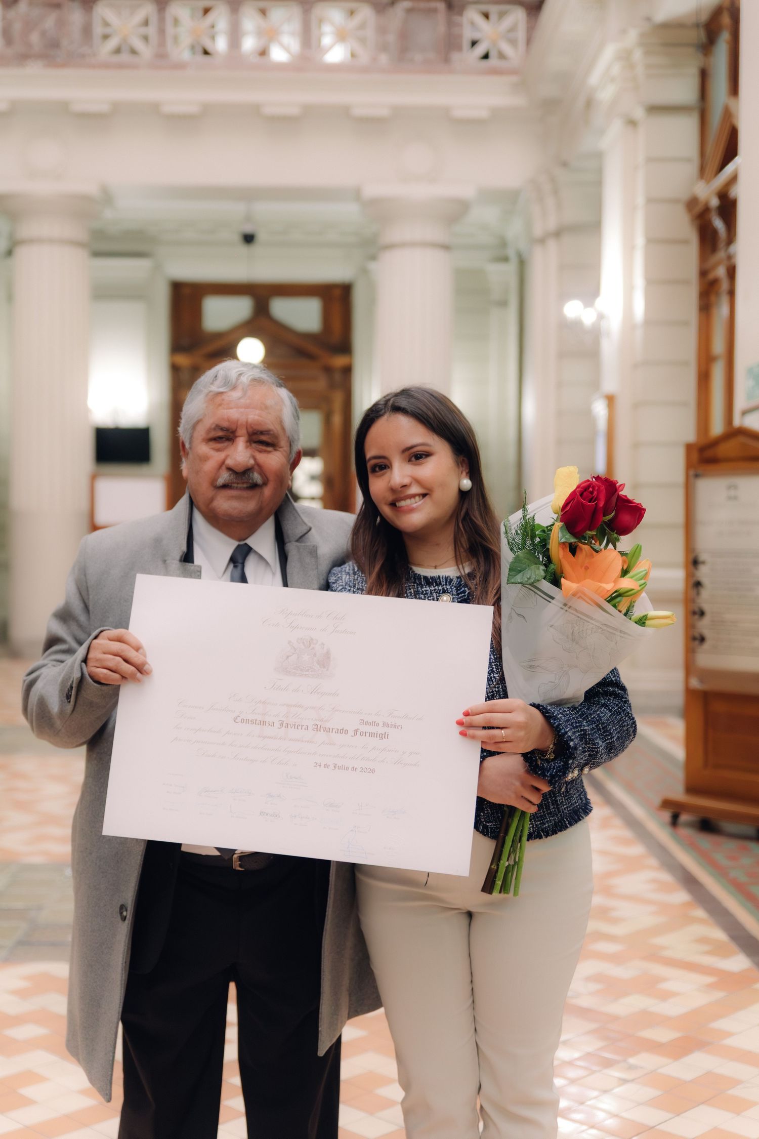 Older gentleman and smiling graduate holding diploma and roses in elegant columned hallway.