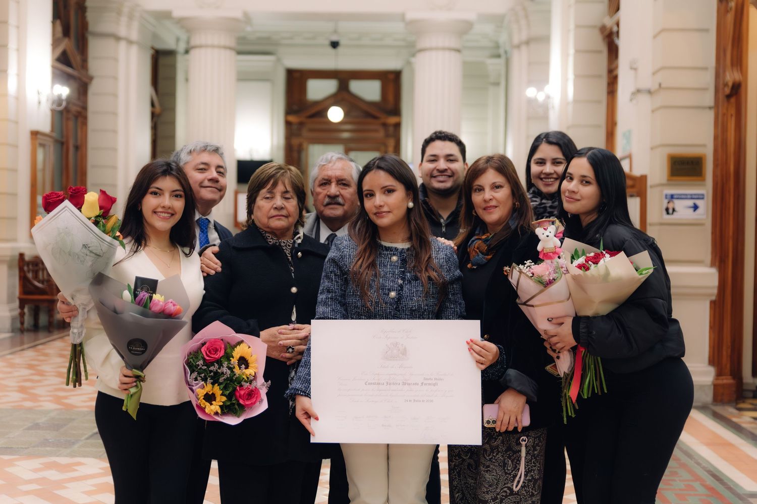 Group of nine people celebrating together indoors holding flowers and an official diploma certificate.