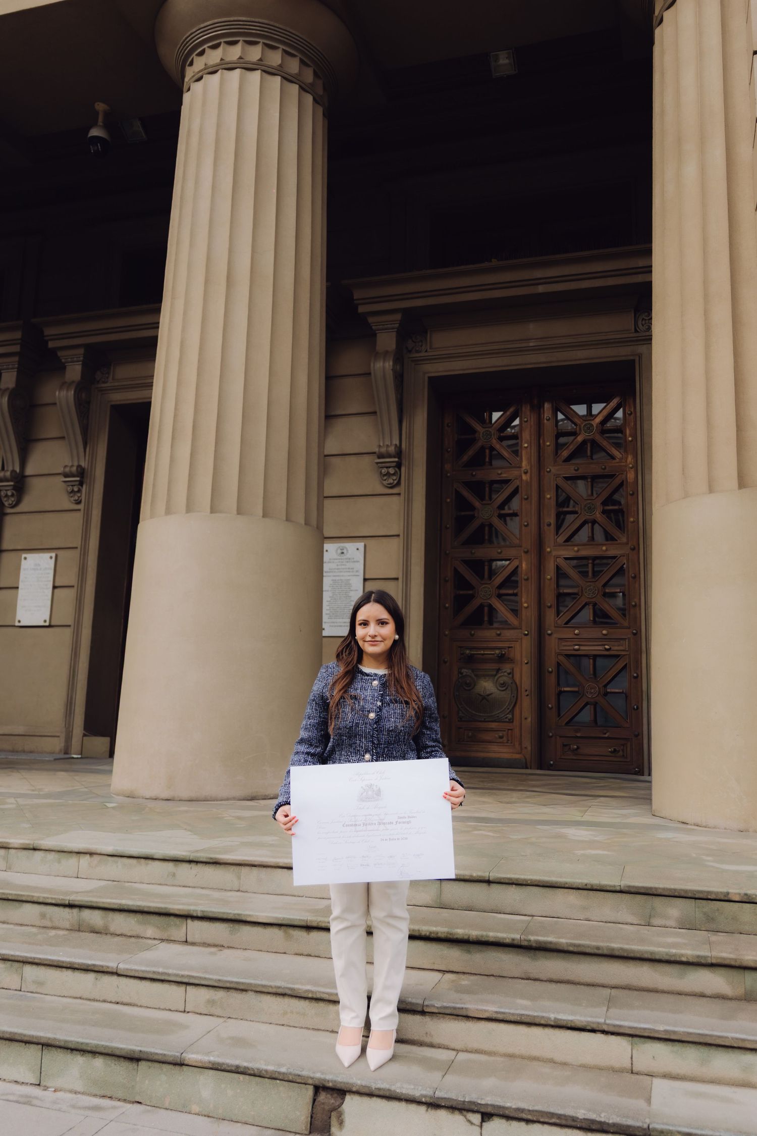 Young woman holding a large official diploma certificate on the steps of a neoclassical building.