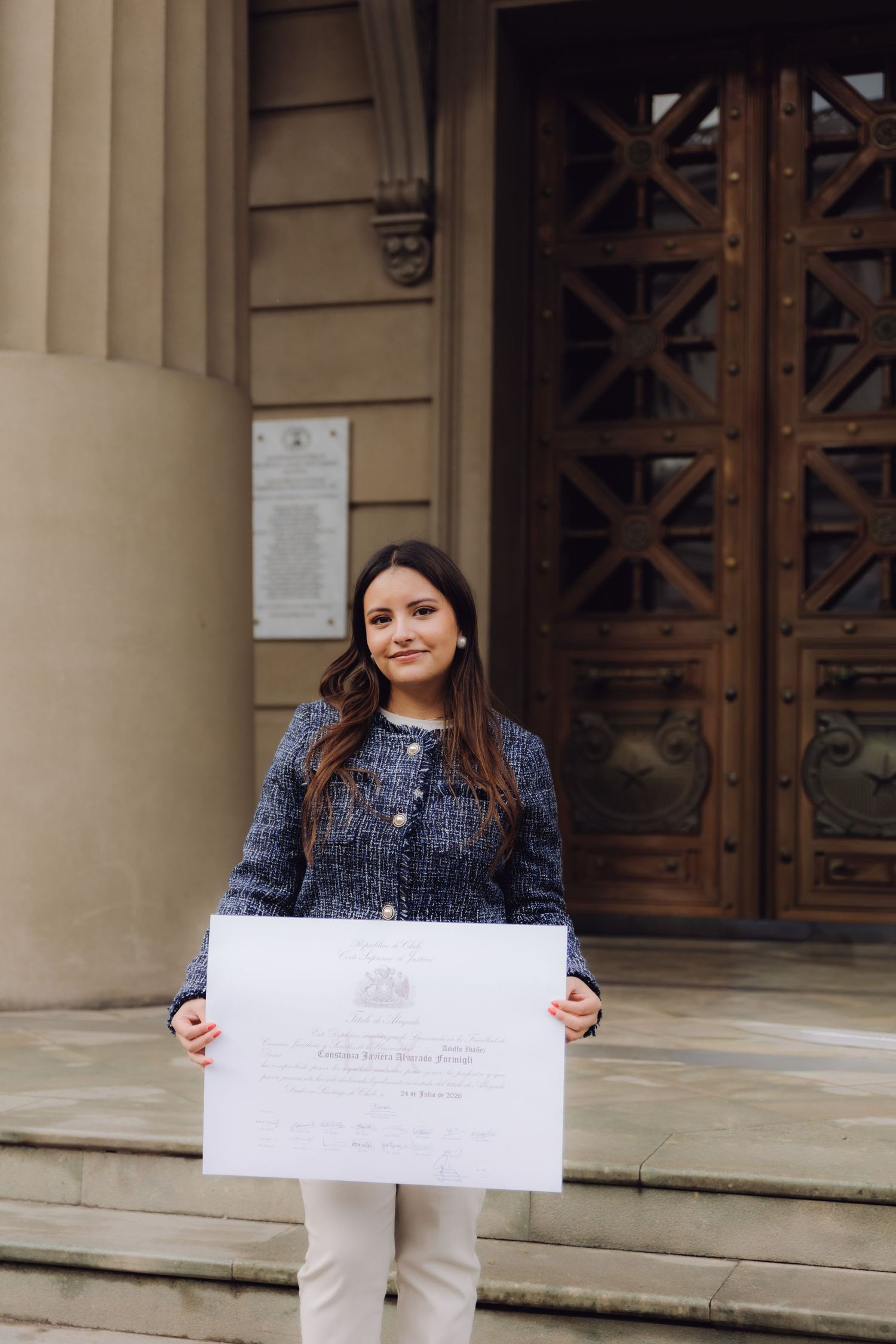 Smiling young woman holding an official diploma certificate outside a historic building with large wooden doors.
