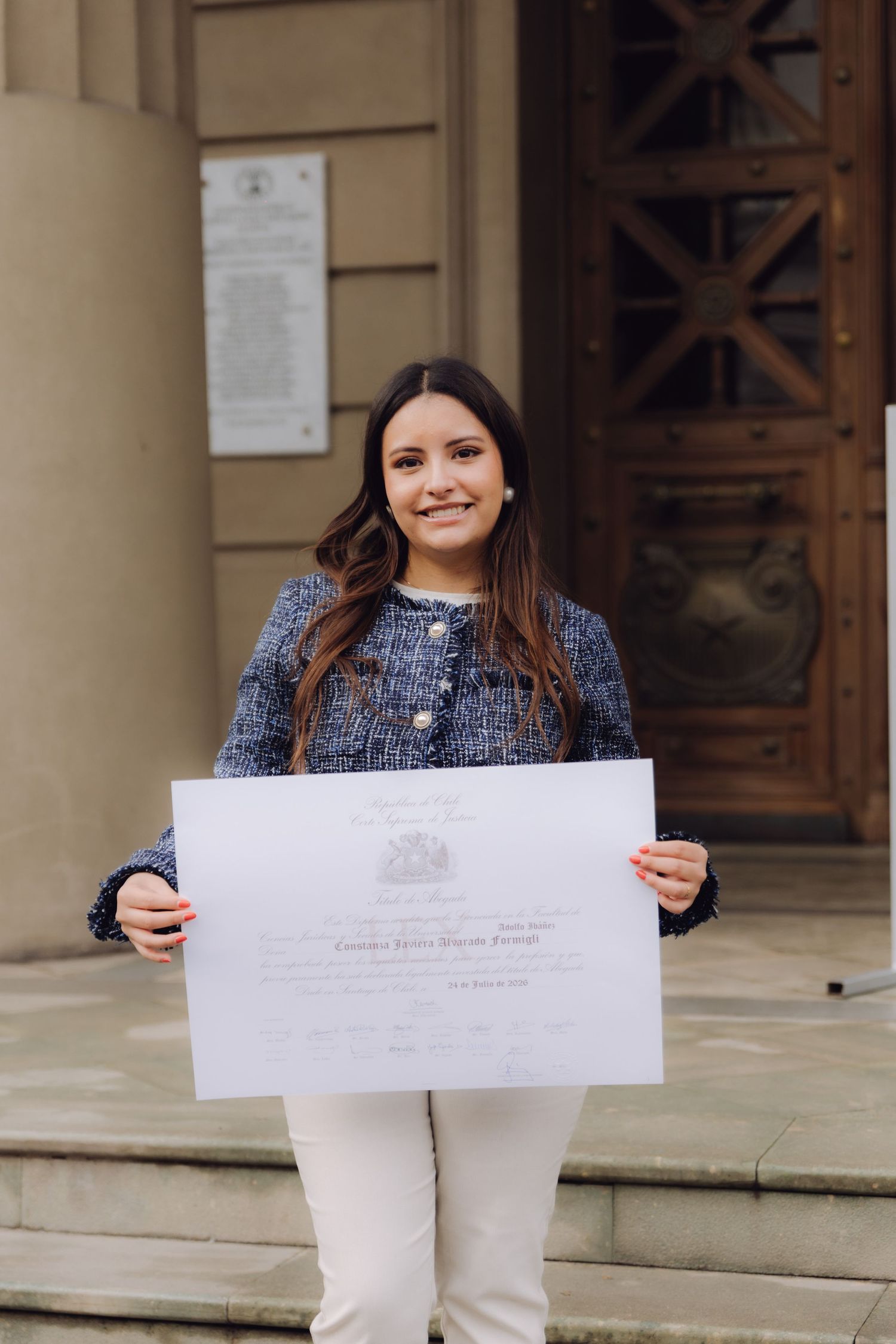 Happy young woman proudly displaying an official diploma certificate on the steps of a grand building.