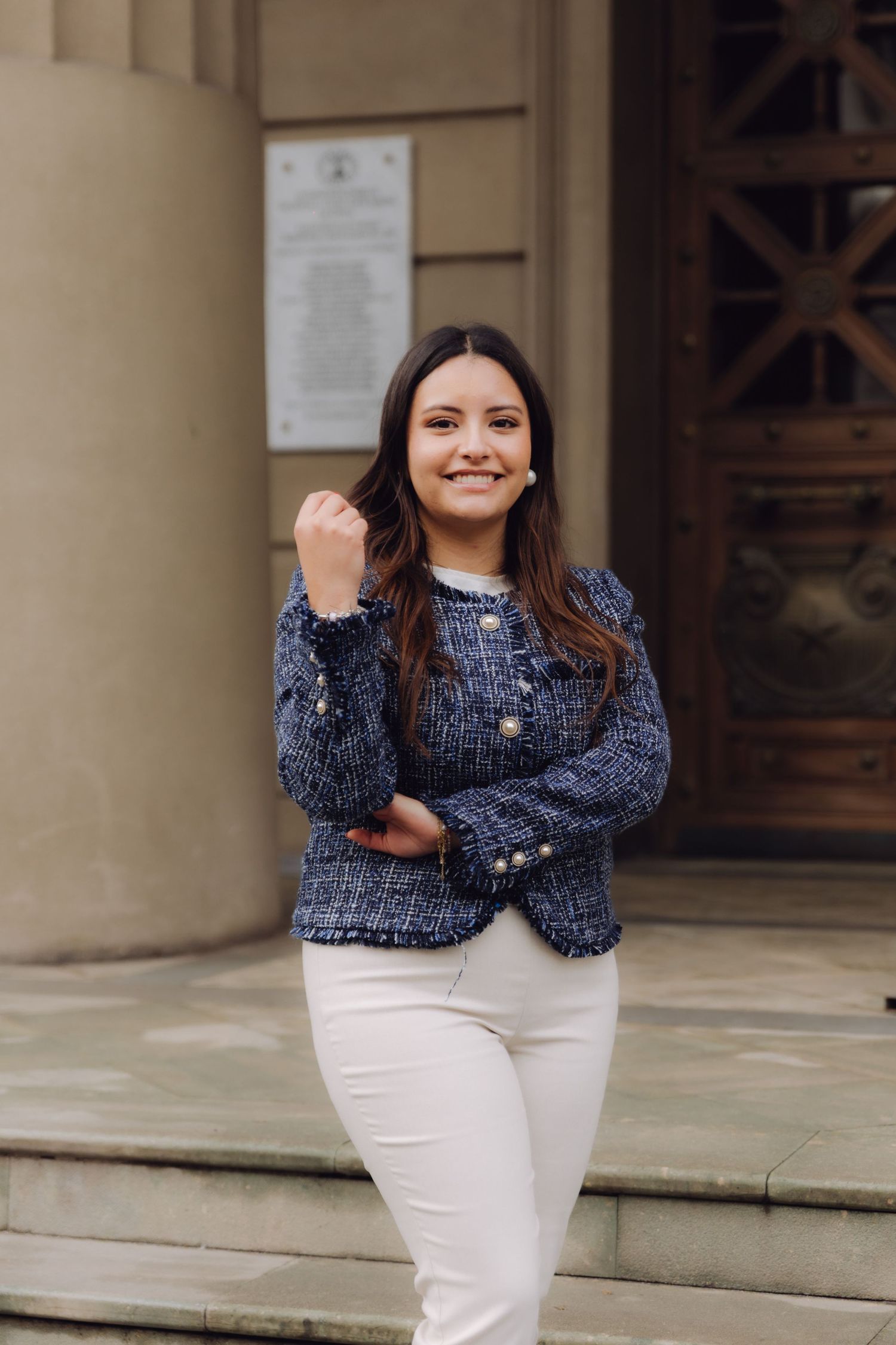 Young woman in blue tweed jacket and white pants smiling confidently on building steps.