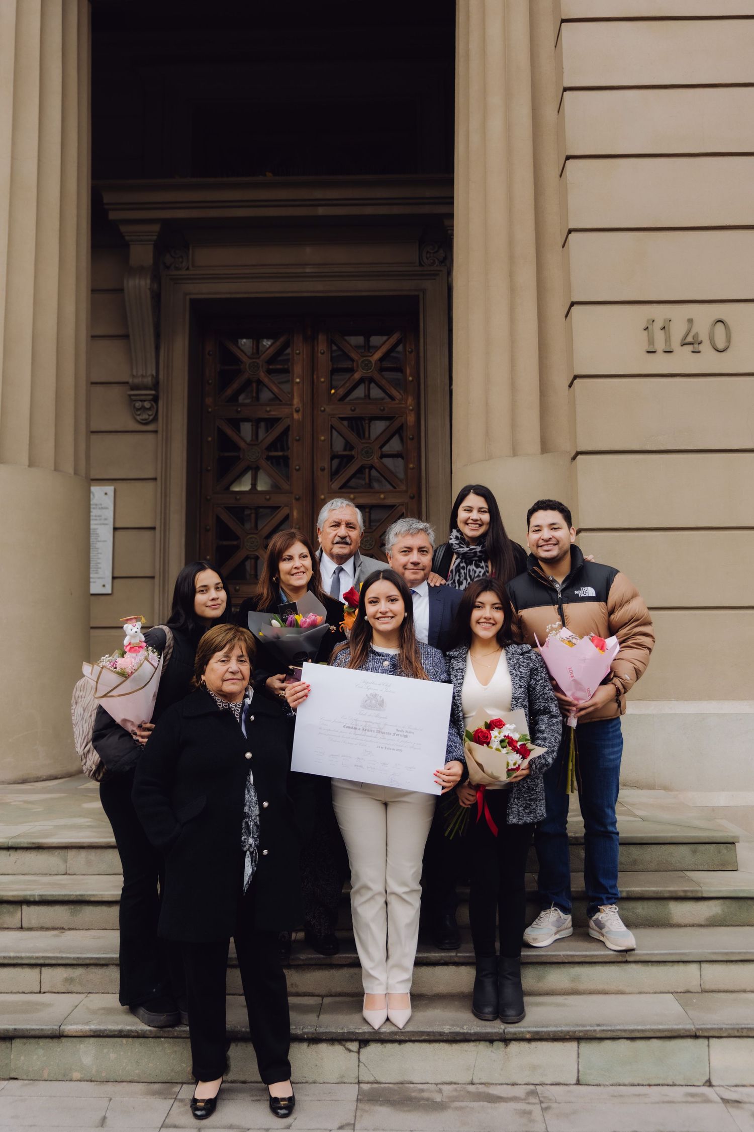 Family group celebrating graduation outdoors on building steps holding diploma and flowers.