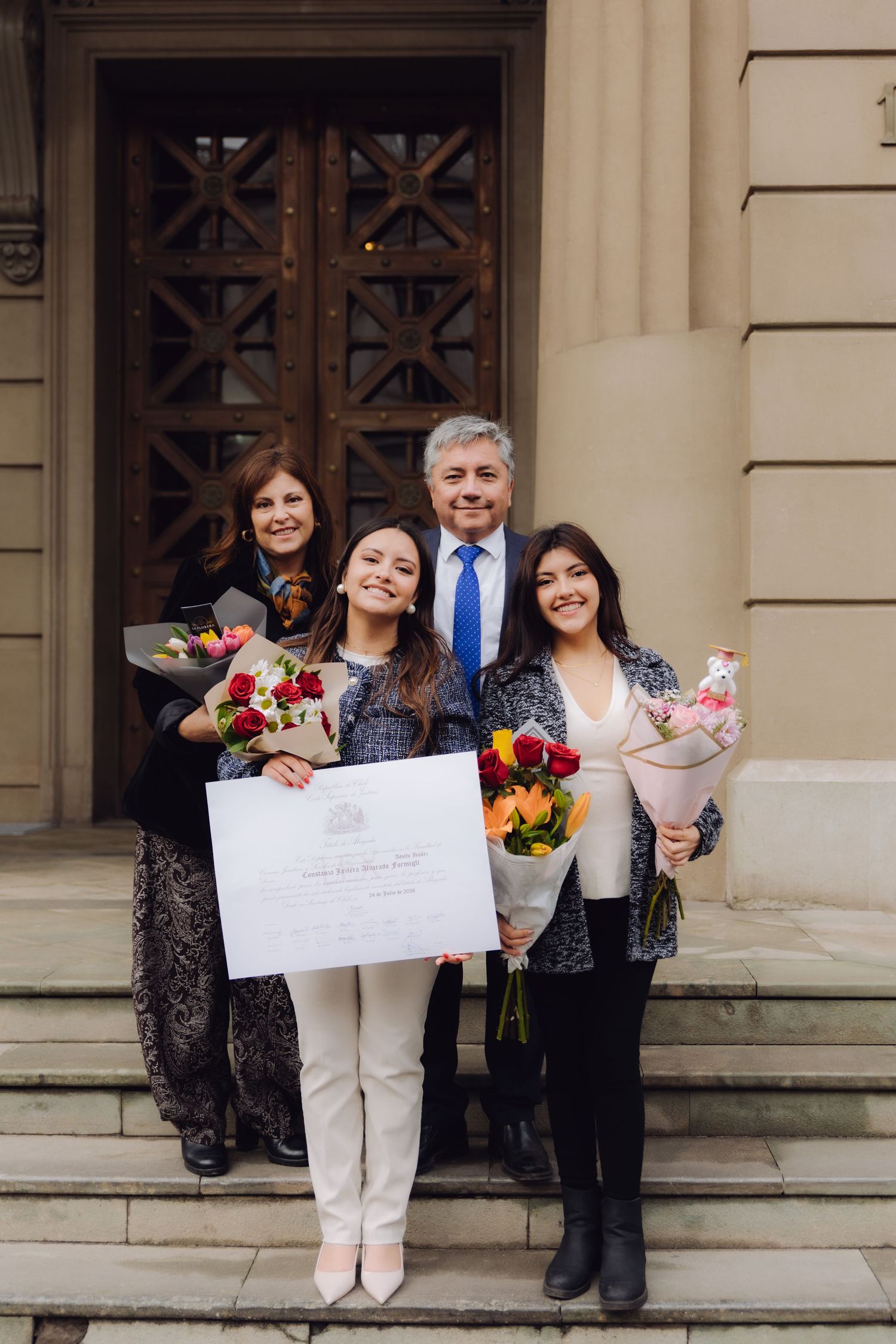 Graduate holding diploma with family holding flower bouquets on grand building steps.