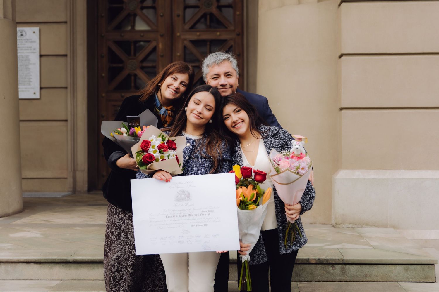 Graduate and family hugging closely on steps holding diploma and colorful flower bouquets.