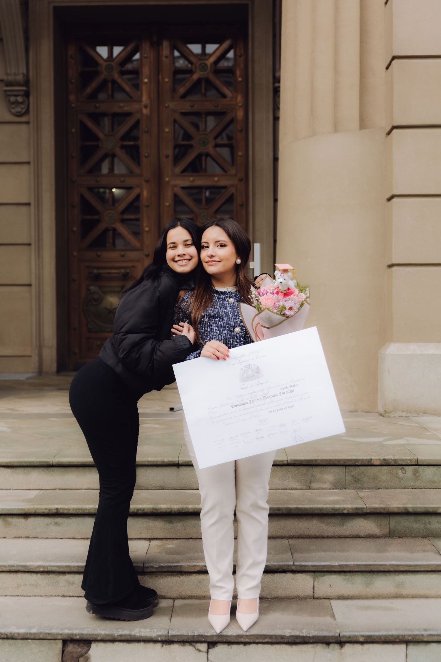 Two young women posing on steps with graduation diploma and pink flower bouquet.