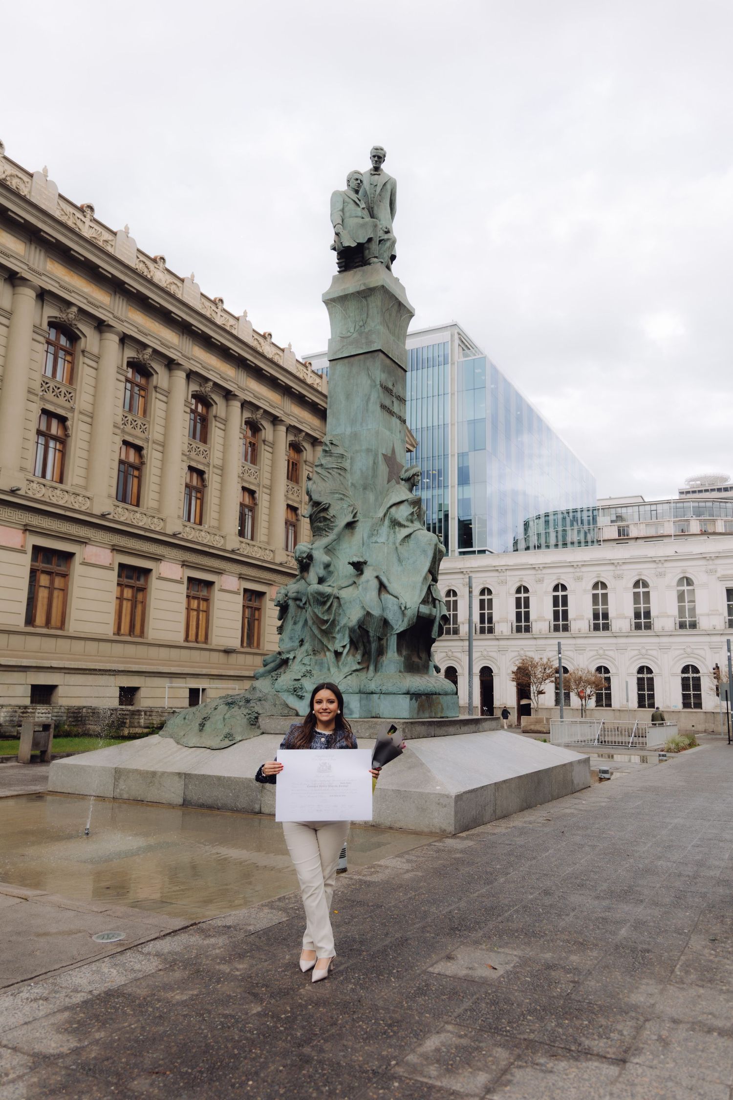 Young woman holding diploma in front of large bronze monument in city plaza.