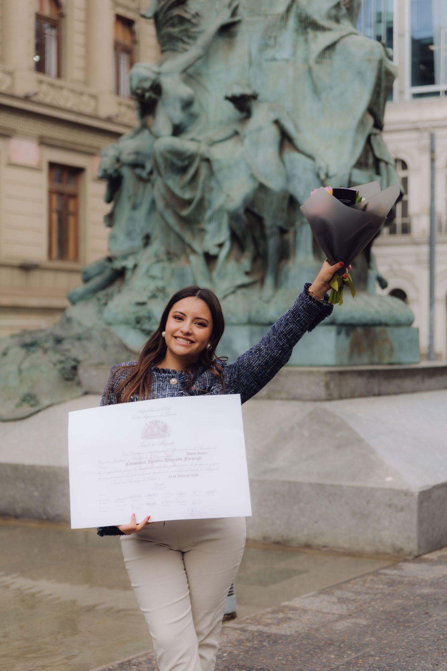 Smiling graduate raising flower bouquet while holding diploma near bronze sculpture outdoors.