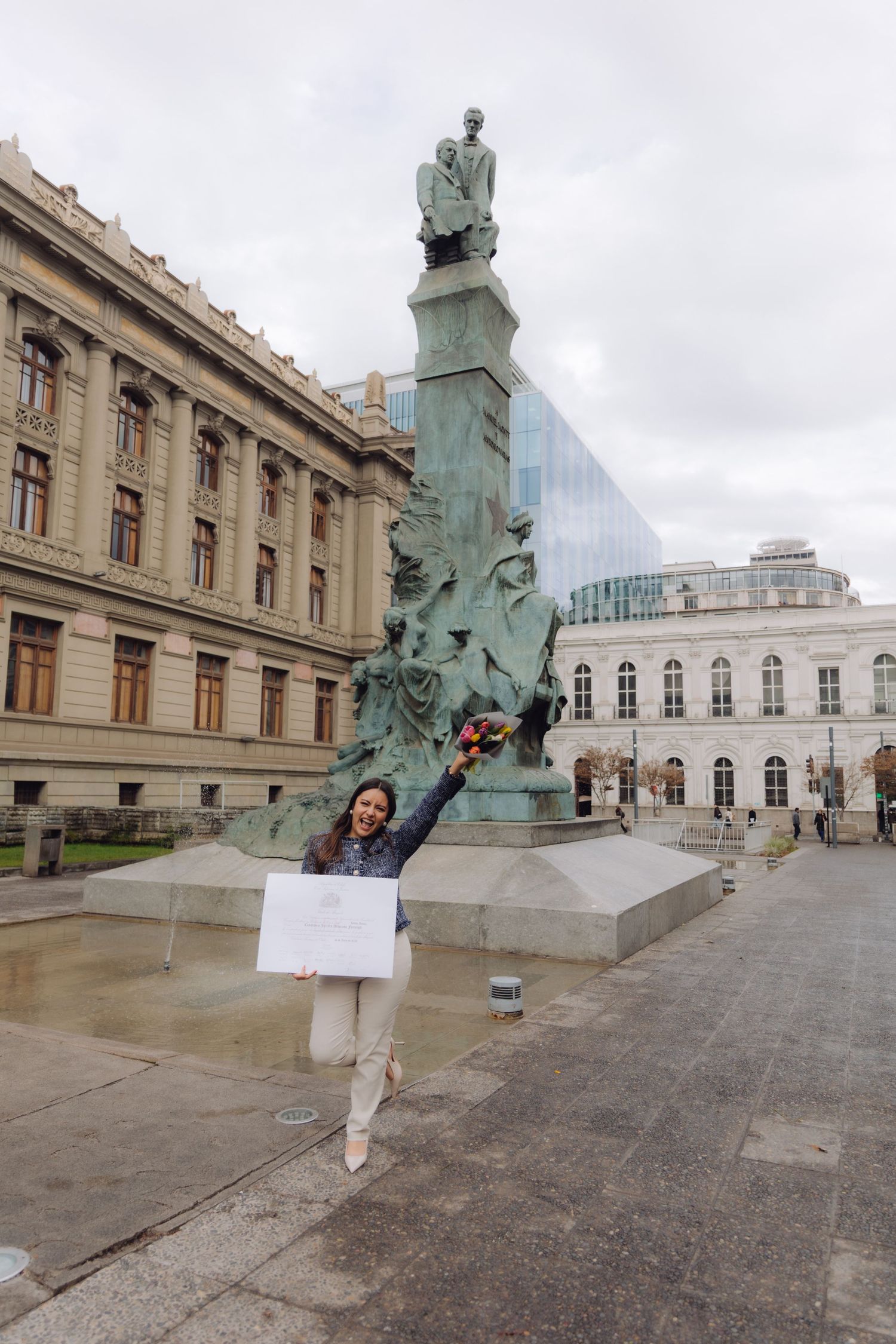 Woman celebrating with raised arm holding flowers and diploma near tall bronze monument.