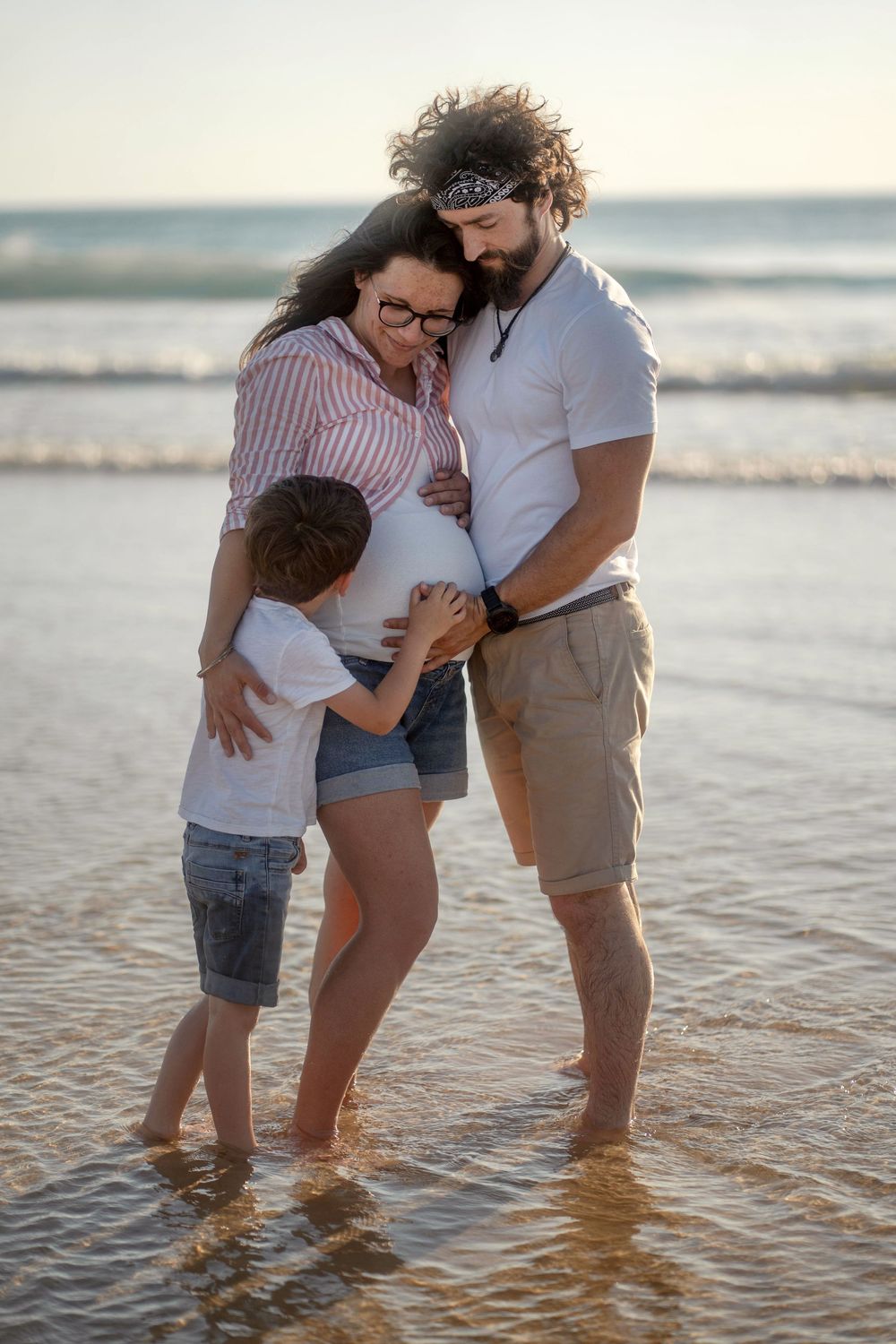 Une famille s'enlacent à la plage, les parents et leur jeune fils touchant le ventre de la future maman au coucher du soleil