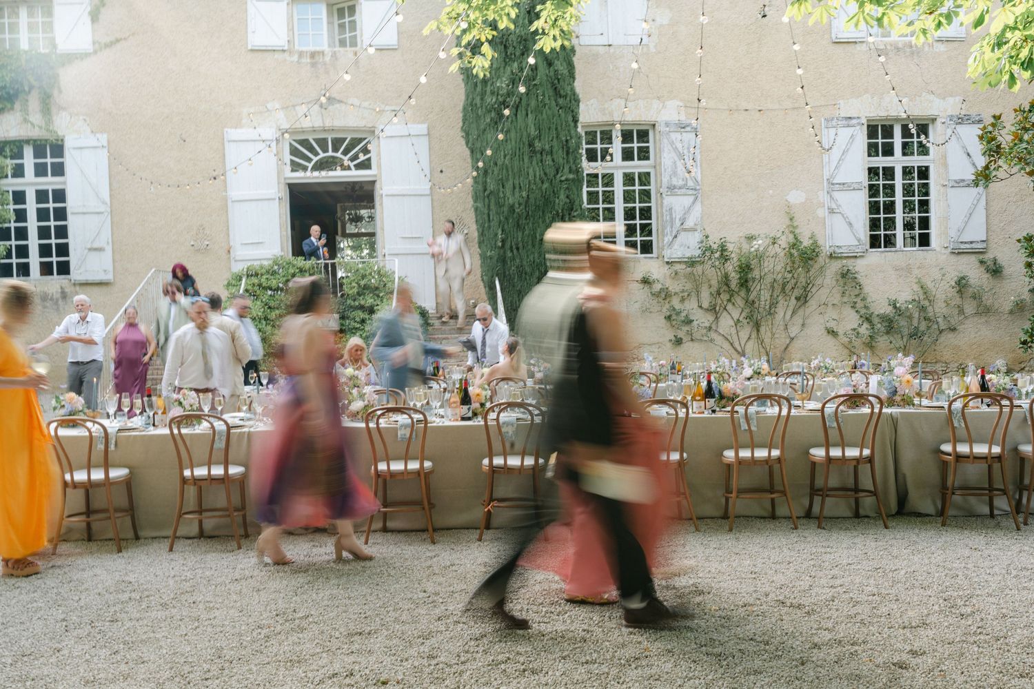 Elegant outdoor wedding reception at a French chateau with long tables, chairs, and blurred guests in colorful attire.