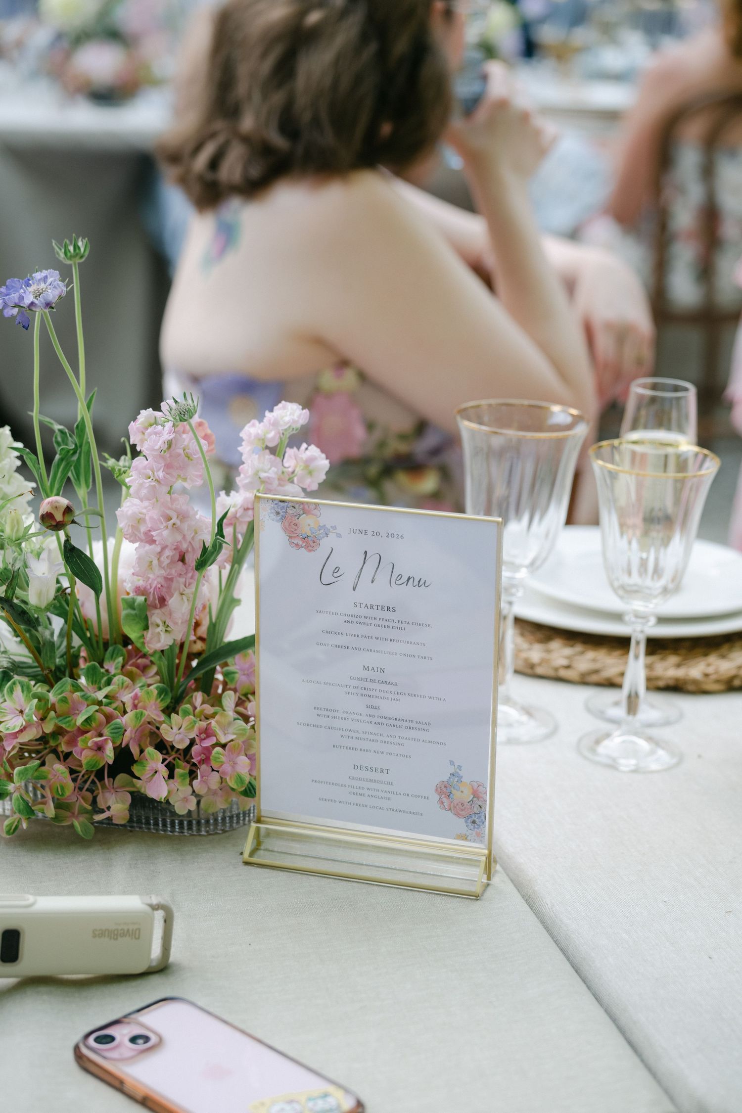 Elegant wedding reception table with floral centerpiece, gold-framed menu card, crystal glasses, and guests in background.