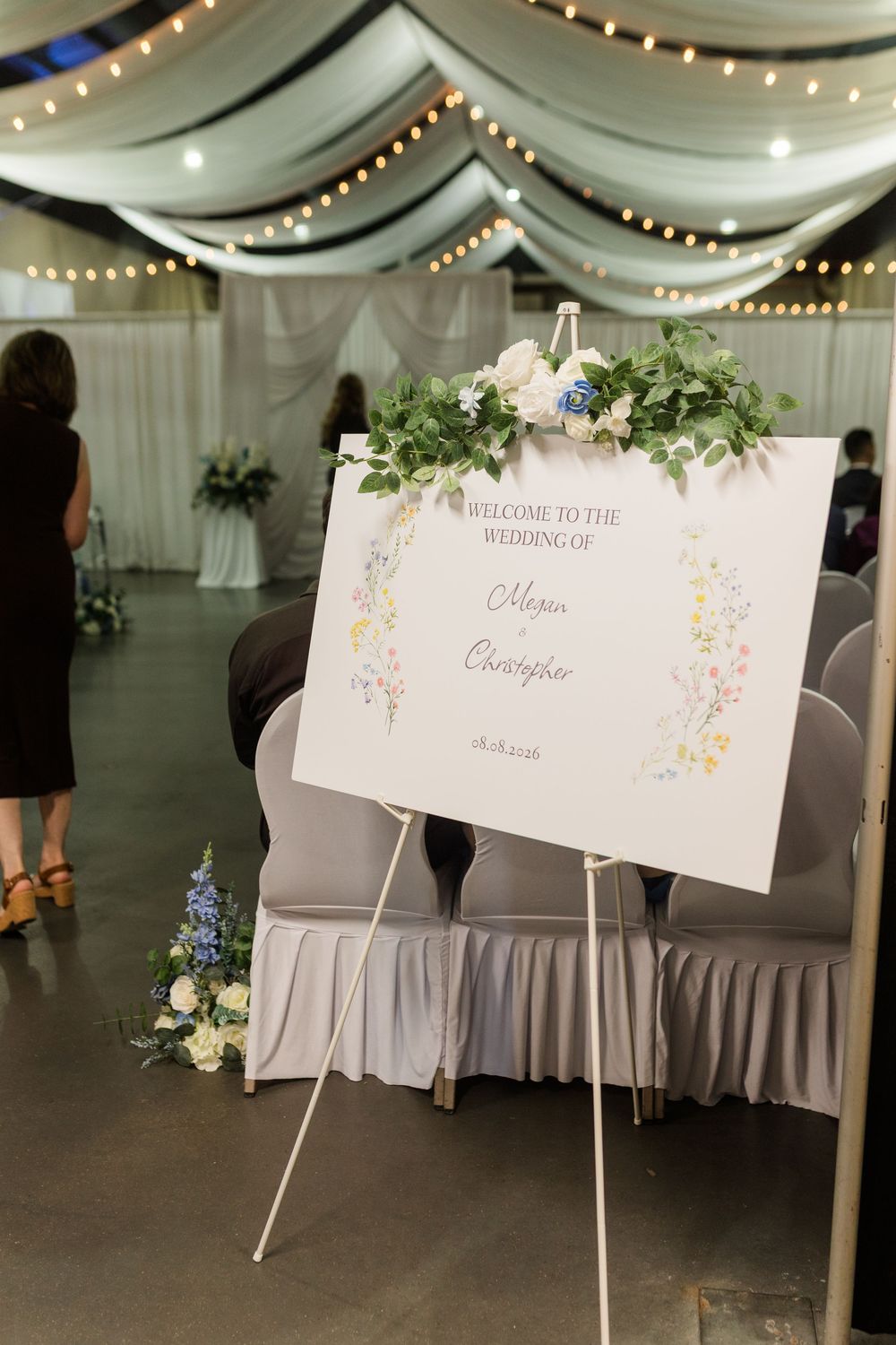 Elegant wedding welcome sign on easel with floral greenery decor in a draped venue with string lights.